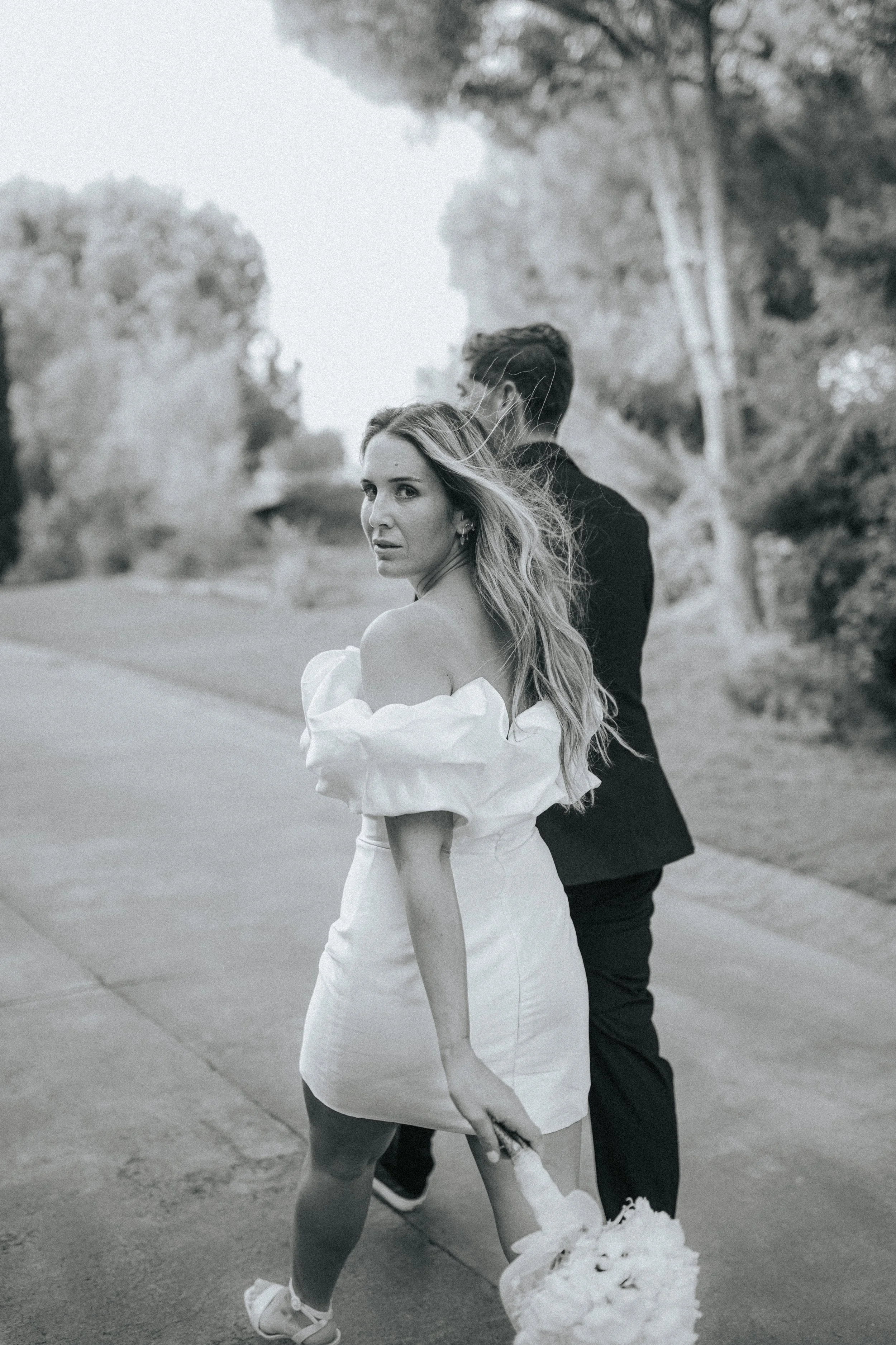 A woman in a white dress holding a bouquet of flowers, with a man standing beside her with his back turned, outdoors on a paved pathway with trees in the background, in black-and-white.