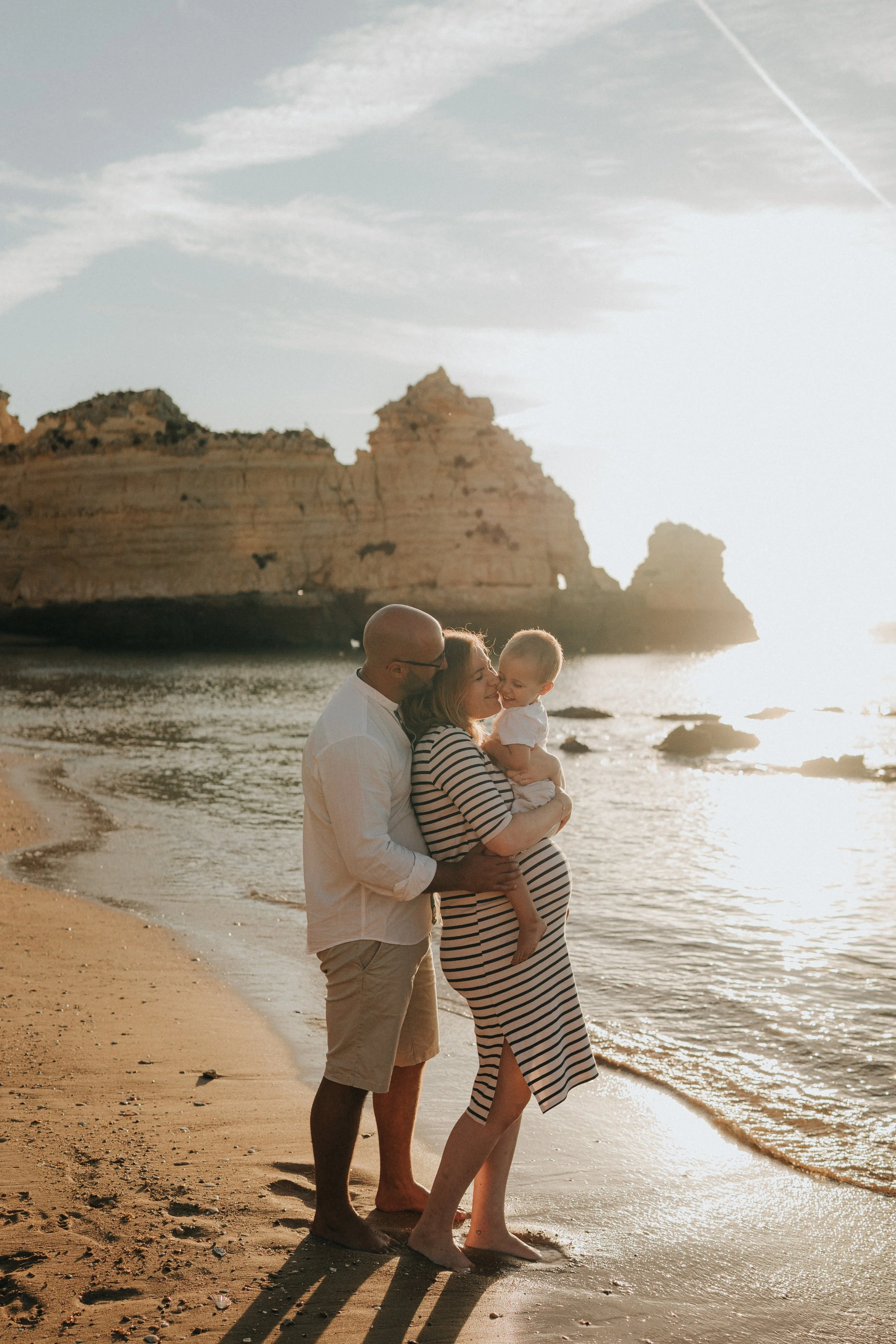 Family of three on the beach, with rocky cliffs in the background, during sunset. The man holds the woman, who is holding a child, both looking at each other.
