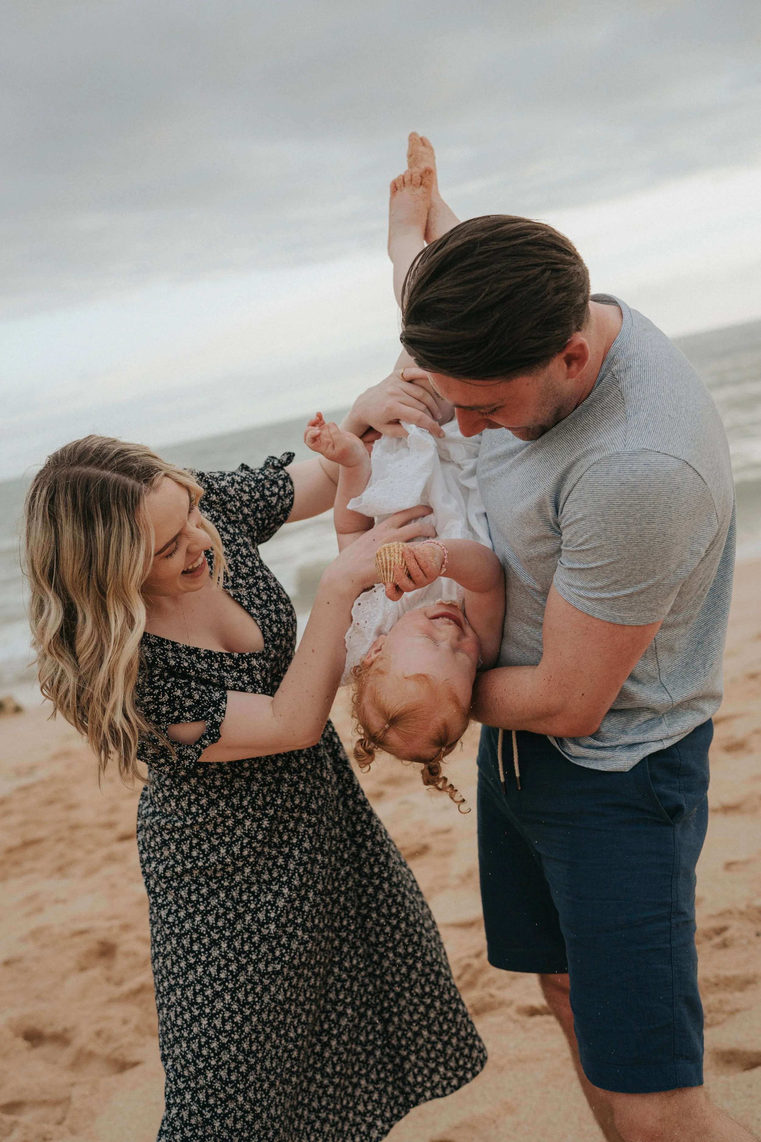 A family at the beach, with a woman holding a child upside down while a man helps hold the child, all smiling and enjoying the moment.