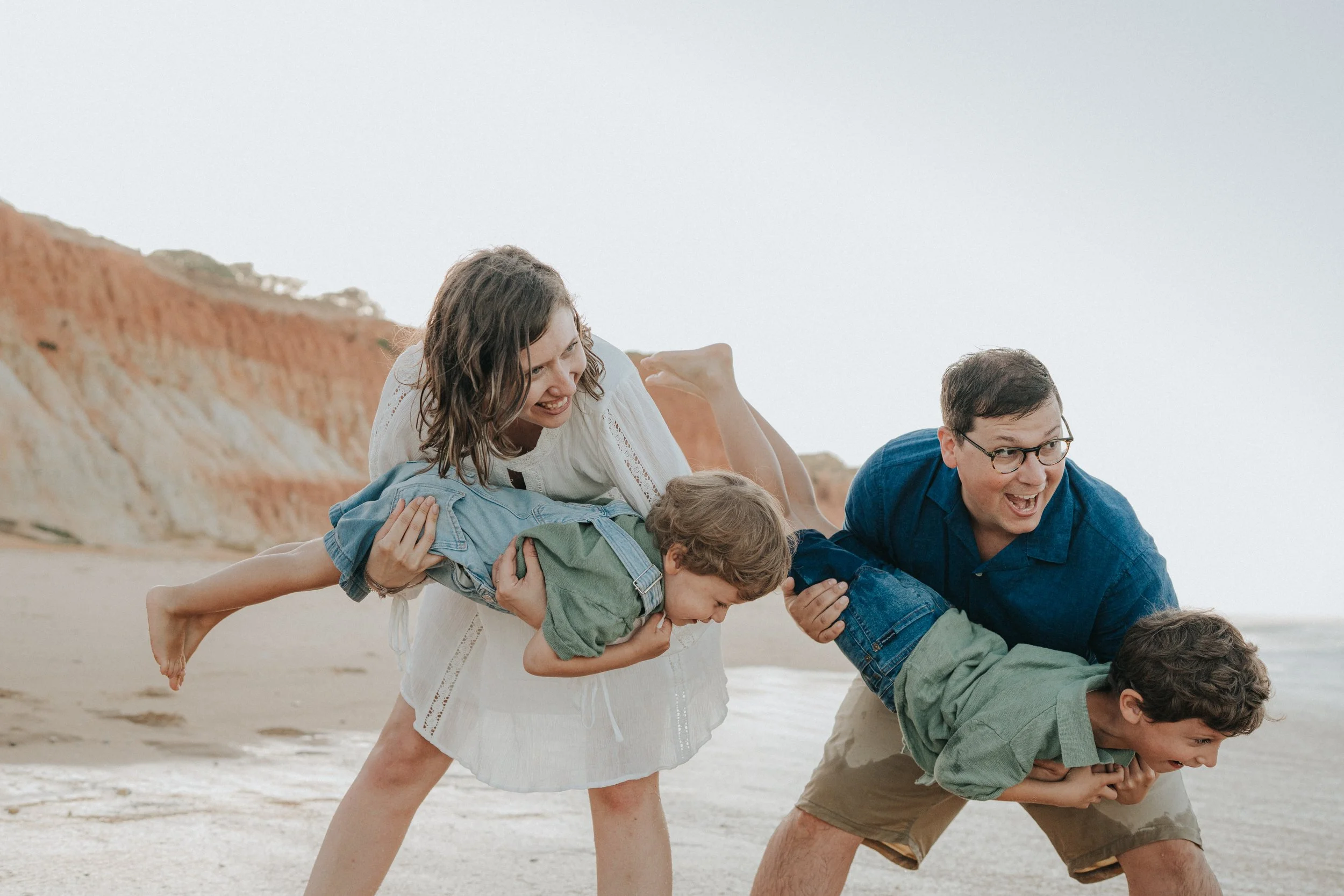 A family of four playing on the beach, forming a human pyramid with the mother at the top, kids in the middle, and the father at the bottom, with cliffs and the ocean in the background.