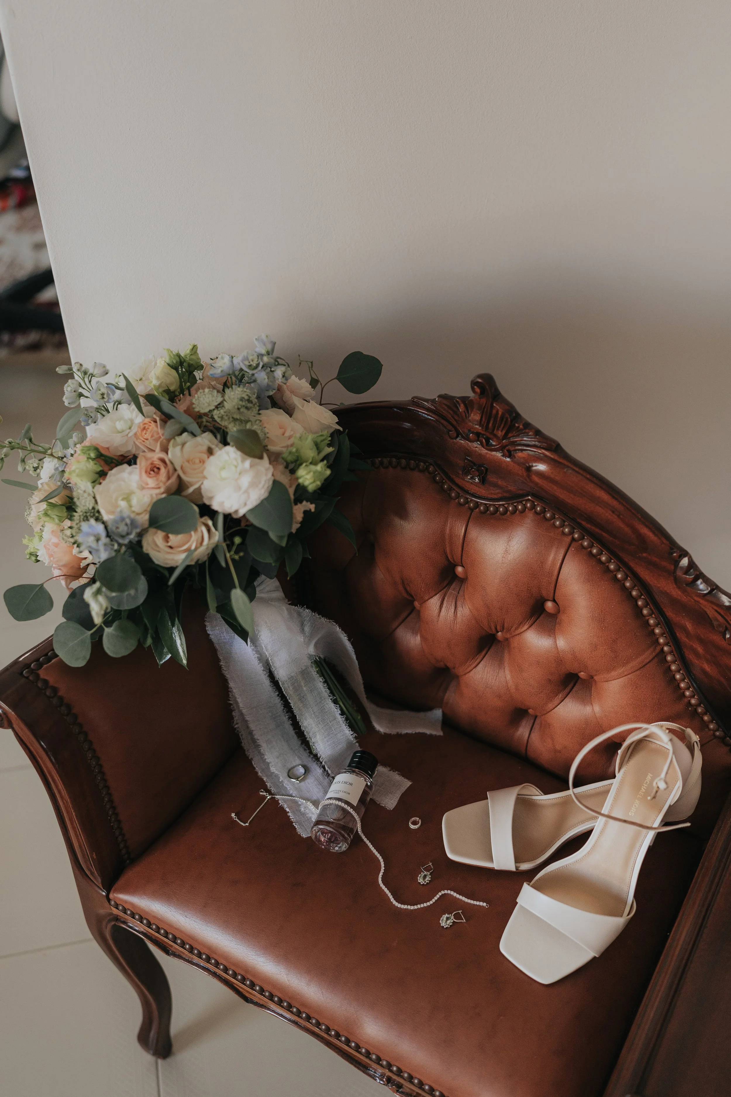 A wooden vintage sofa with a floral bouquet, perfume bottle, jewelry, and a pair of white high-heeled shoes on it.