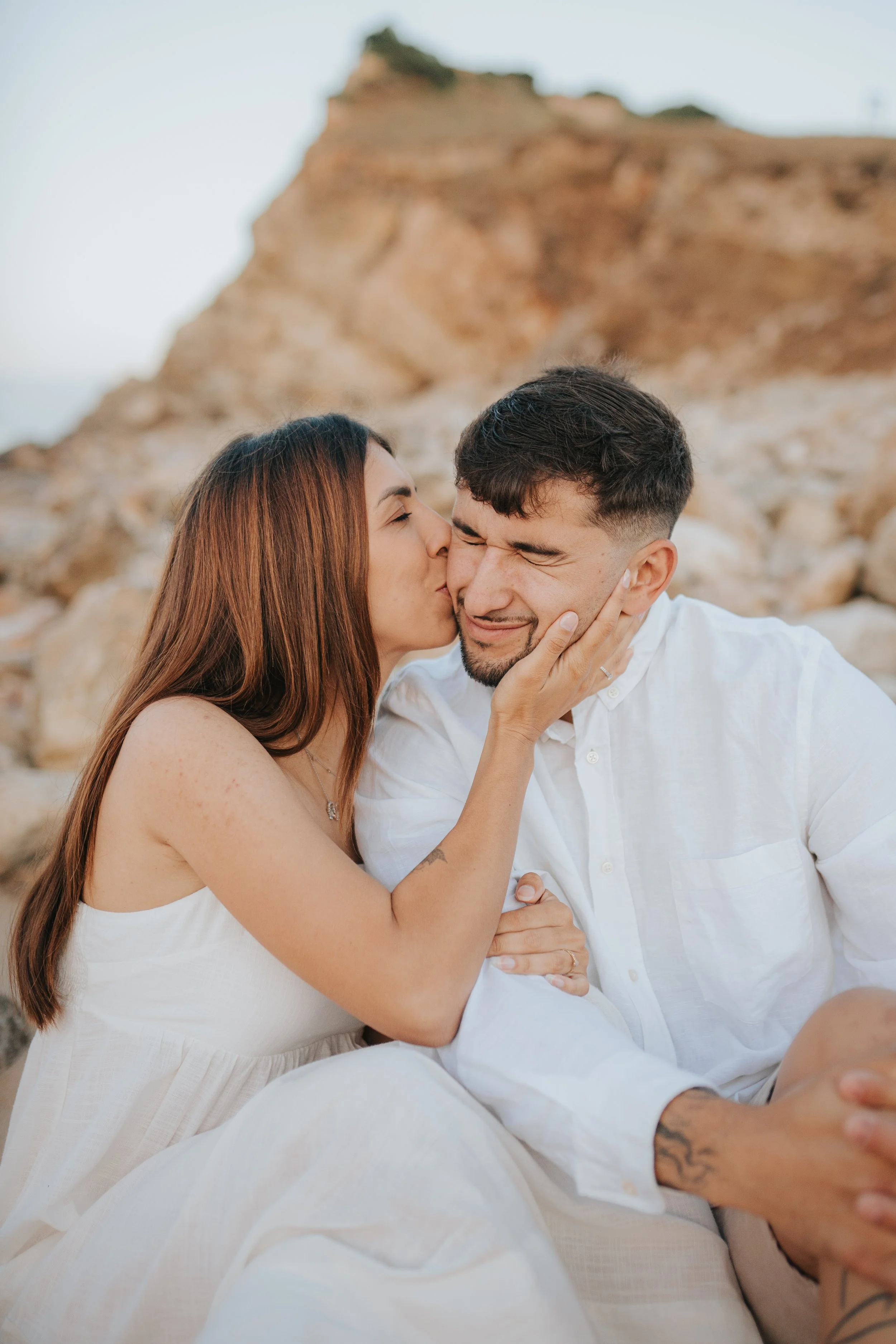 A couple sitting close together outdoors, with the woman kissing the man on the cheek, both smiling and embracing each other against a rocky background.