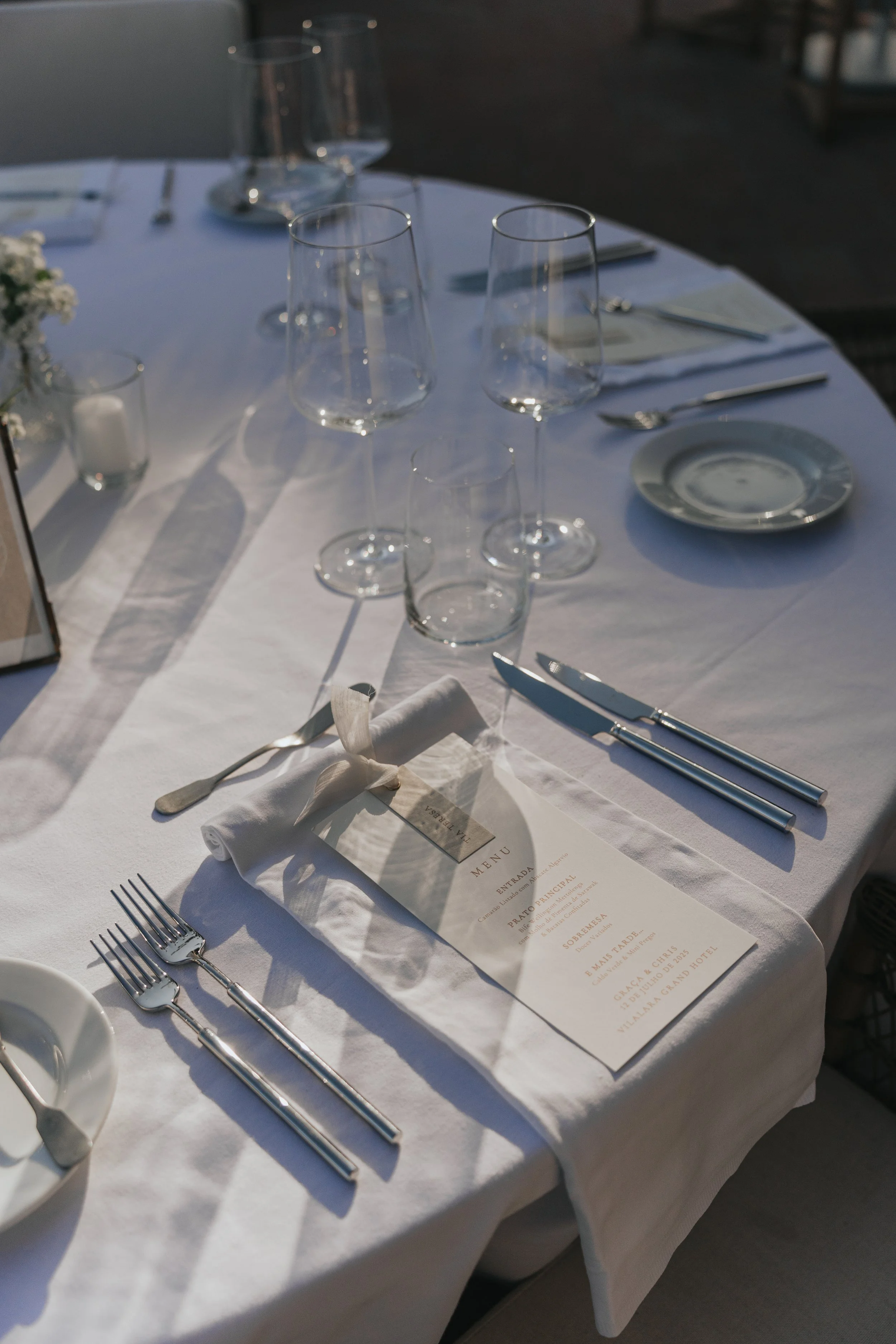 Table set for a formal dining event with wine glasses, plates, silverware, and a menu, all arranged on a white tablecloth.