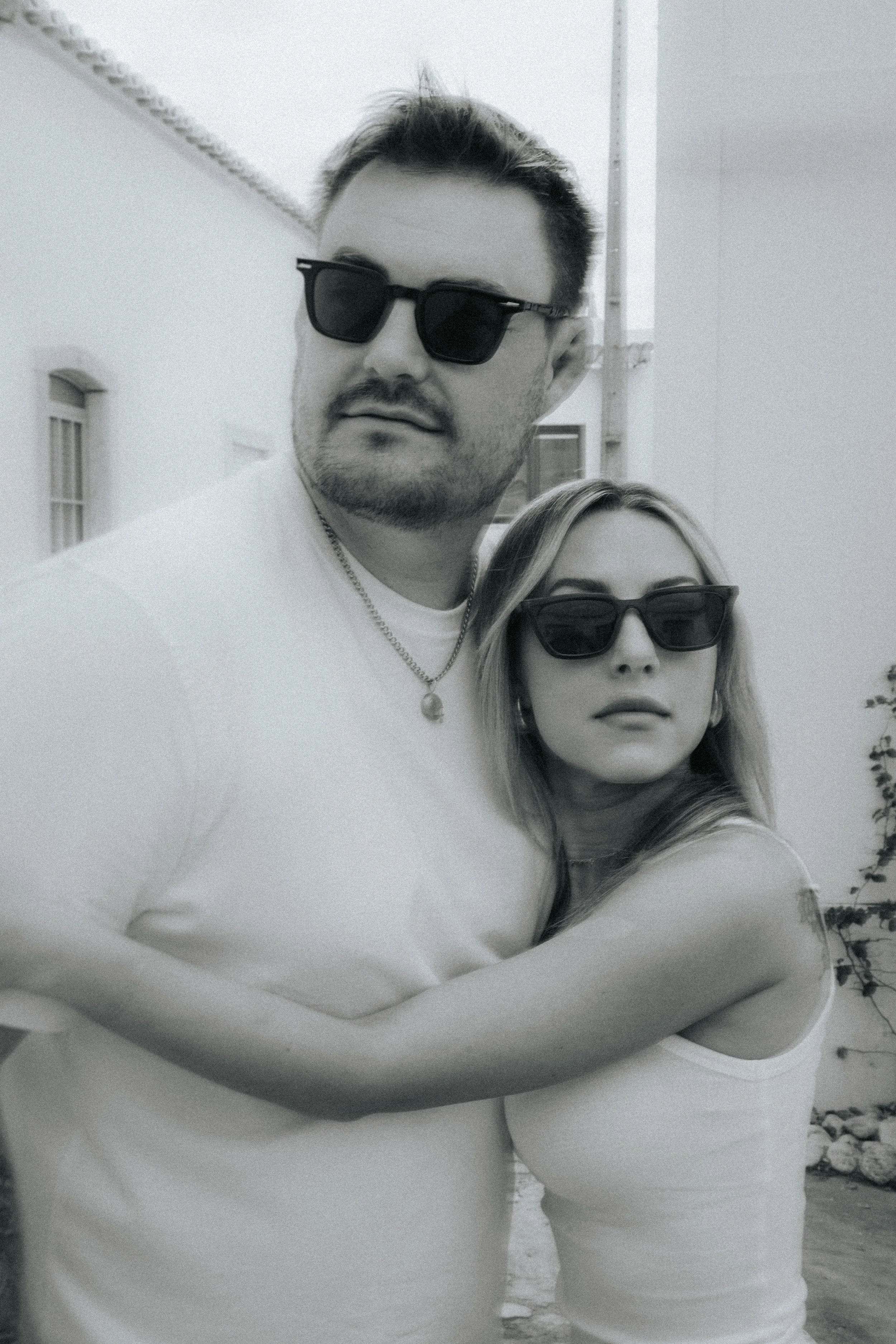 A man and a woman both wearing sunglasses, standing outside on a bright day. The man has short hair, a beard, and is wearing a white shirt and a necklace. The woman has long hair and is wearing a white top.