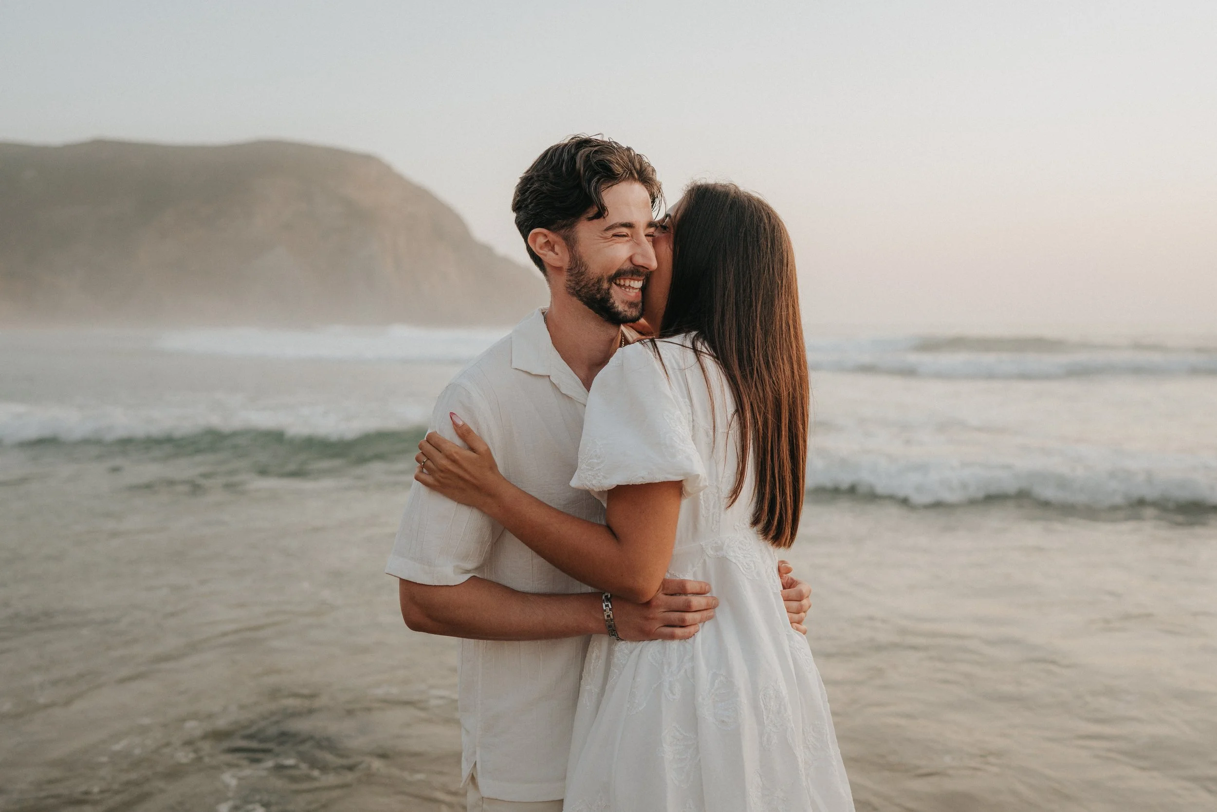 A couple hugging on the beach, with the woman kissing the man's cheek, both smiling. The man has dark hair and facial hair, and the woman has long brown hair. The background shows a sandy beach with gentle waves and a distant hilly landscape.