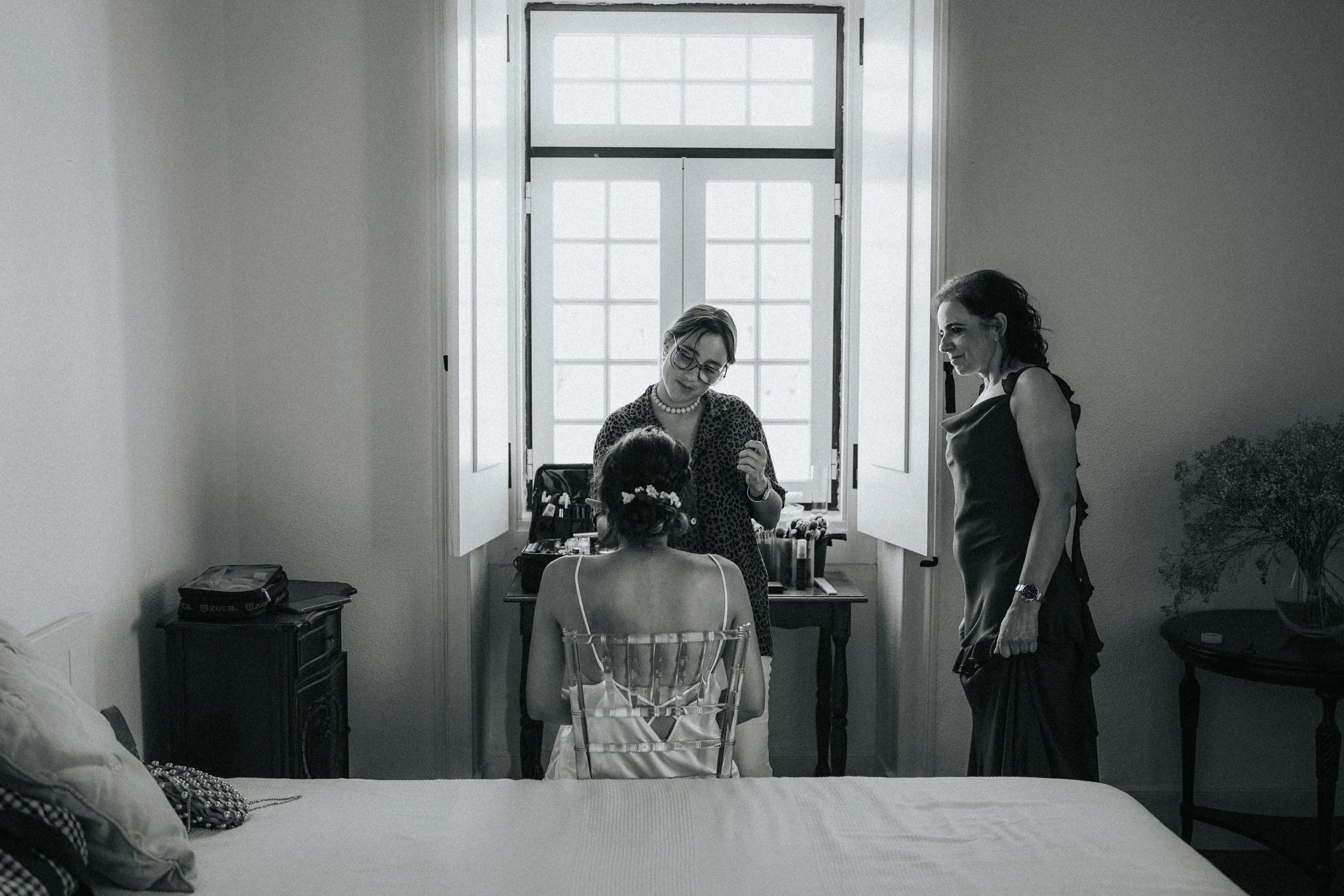 A woman is getting her hair styled by a hairstylist in a room with large windows. There is a woman sitting in a transparent chair with her back to the camera, and two other women, one with glasses and a pearl necklace, are smiling and talking. The ro