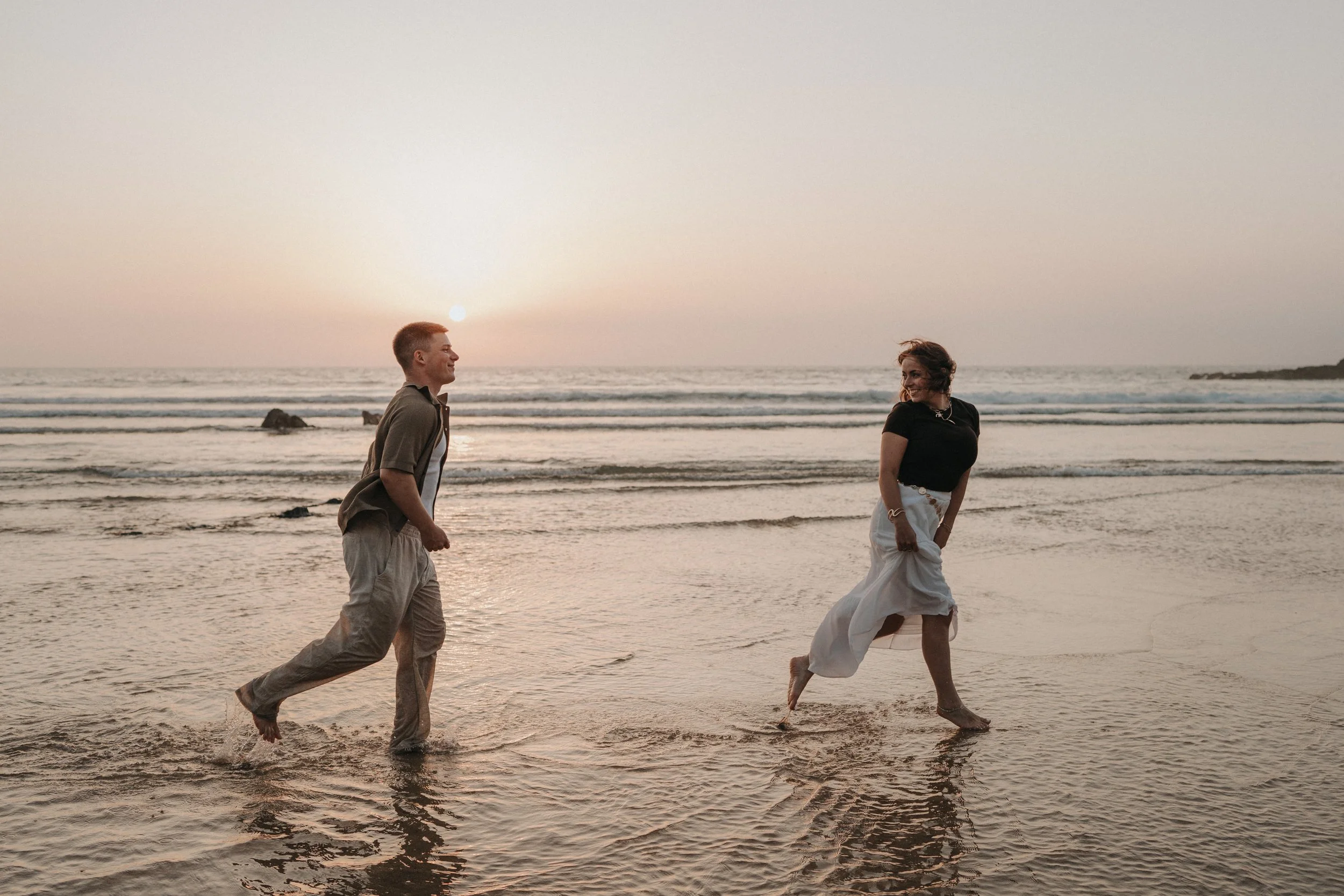 A man and woman walk on the beach during sunset, smiling and holding hands, with waves and rocks in the background.