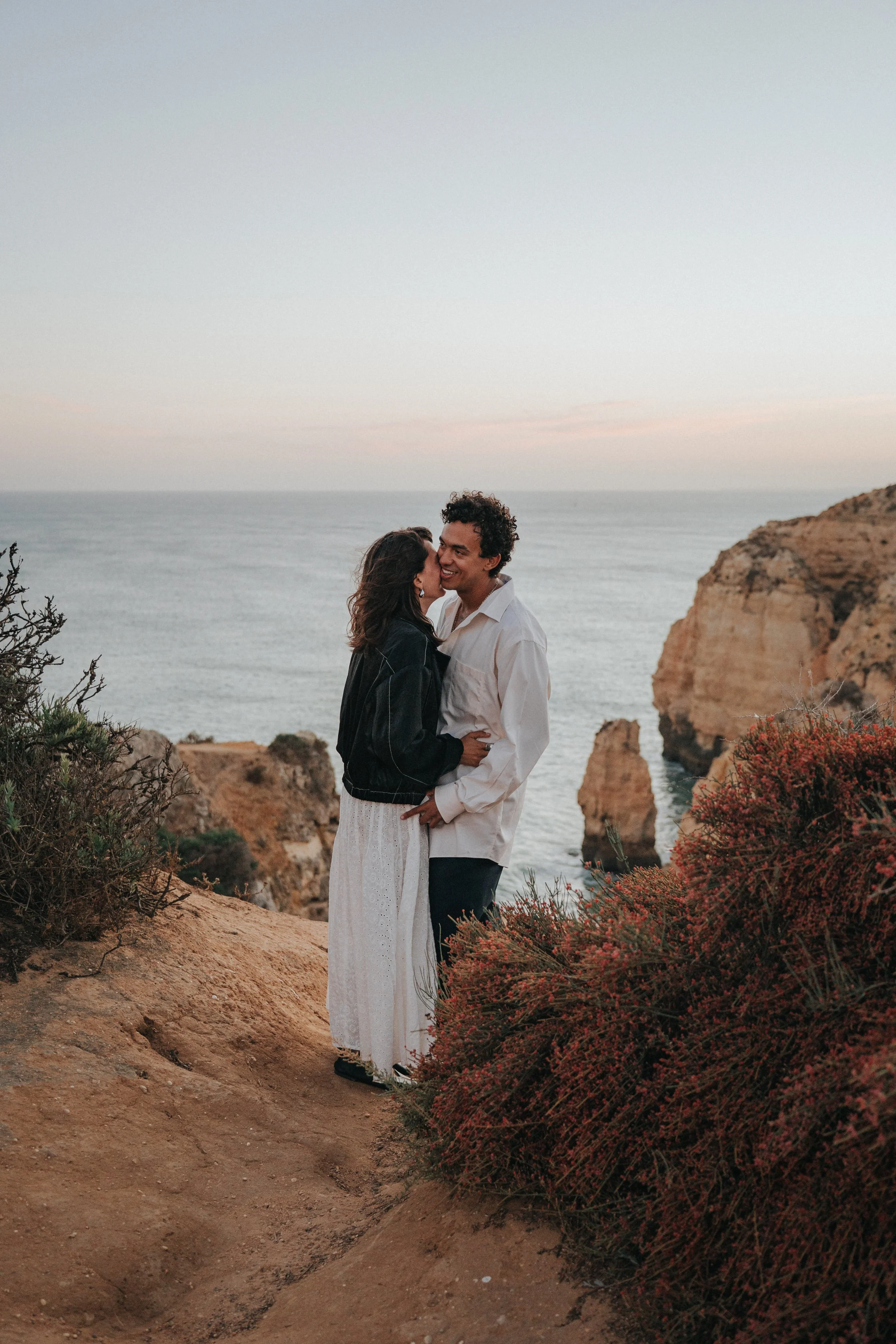 A couple embracing on a rocky cliff near the ocean at sunset, with scenic cliffs and water in the background.