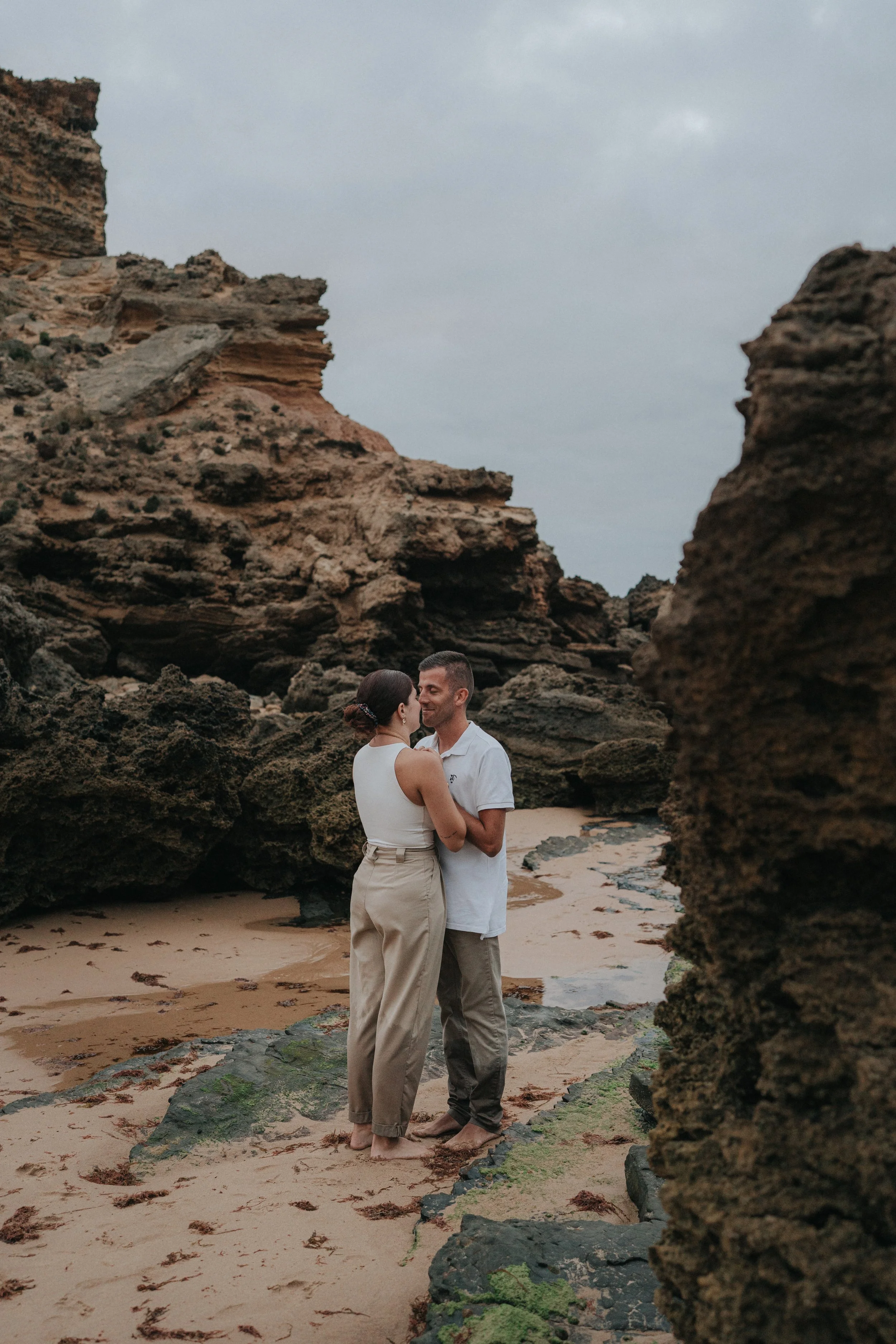 A couple is standing close together and looking into each other's eyes on a sandy beach with large rocks and cliffs around them.