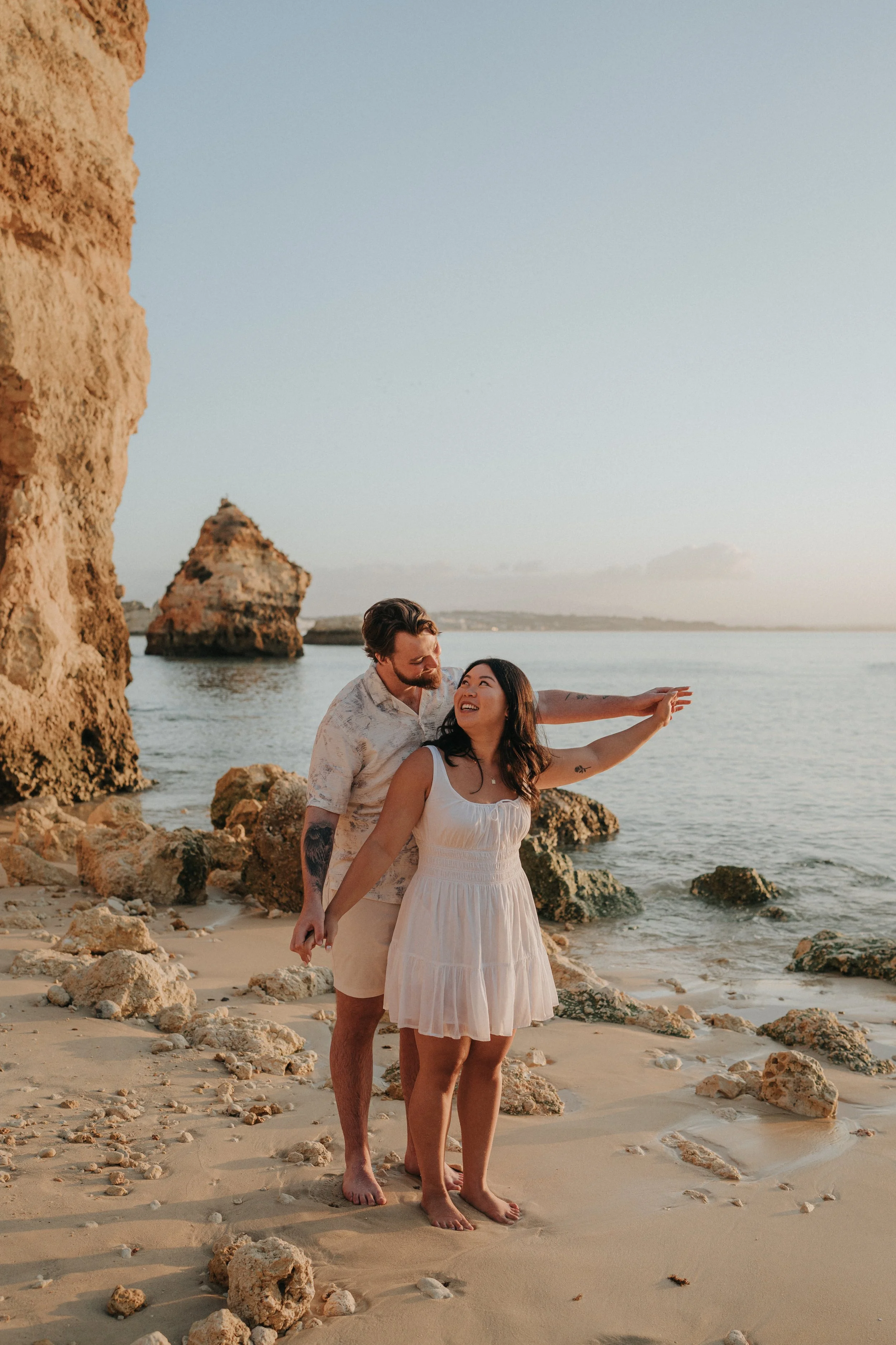 A young couple standing on a rocky beach during sunset, with the man wrapping his arms around the woman, who is smiling and pointing towards the distance, near the ocean with rocks and cliffs in the background.