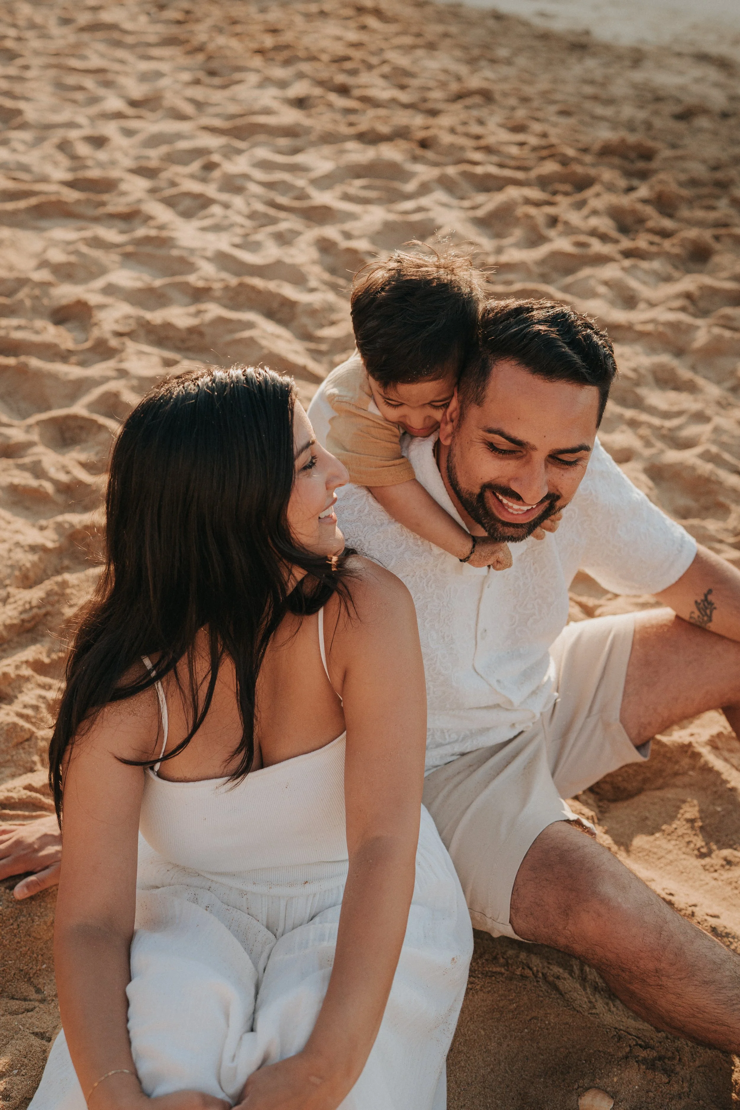 A happy family sitting on the sand at the beach, with a woman, a man, and a young boy, all smiling and enjoying the sunny day.