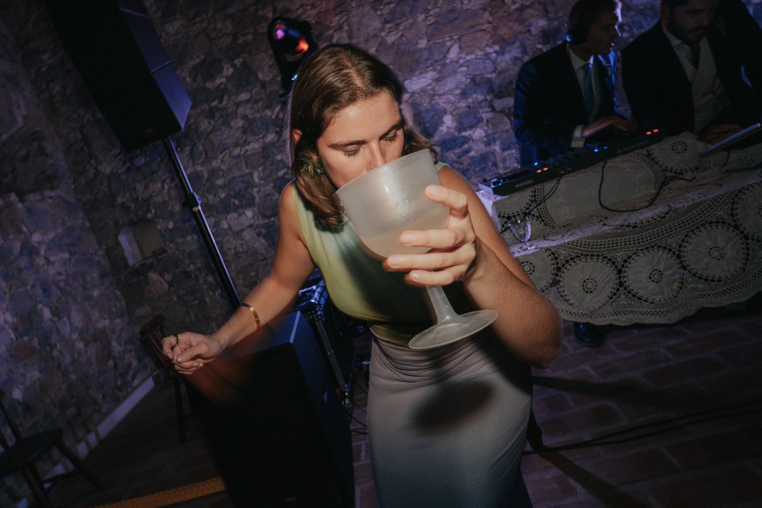 Woman drinking from a large glass at a party, with DJ playing music in the background.