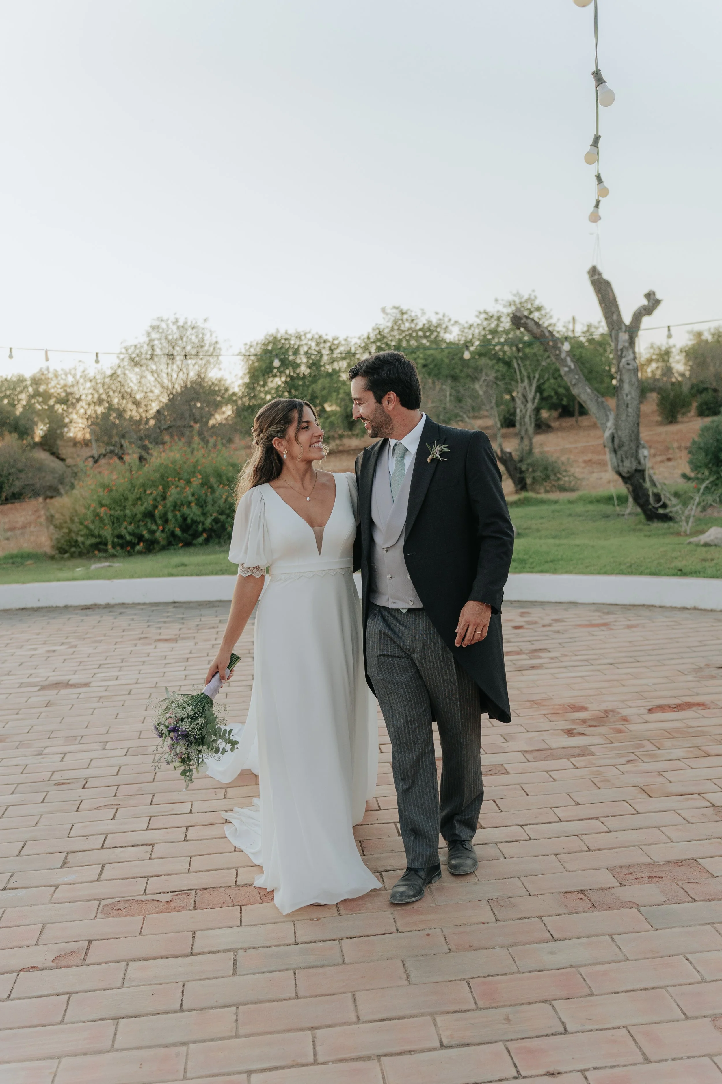 A bride and groom walking outdoors on a brick-paved area, smiling at each other. The bride is holding a bouquet of flowers and wearing a white wedding dress, while the groom is wearing a dark suit with a boutonniere. There are trees and string lights