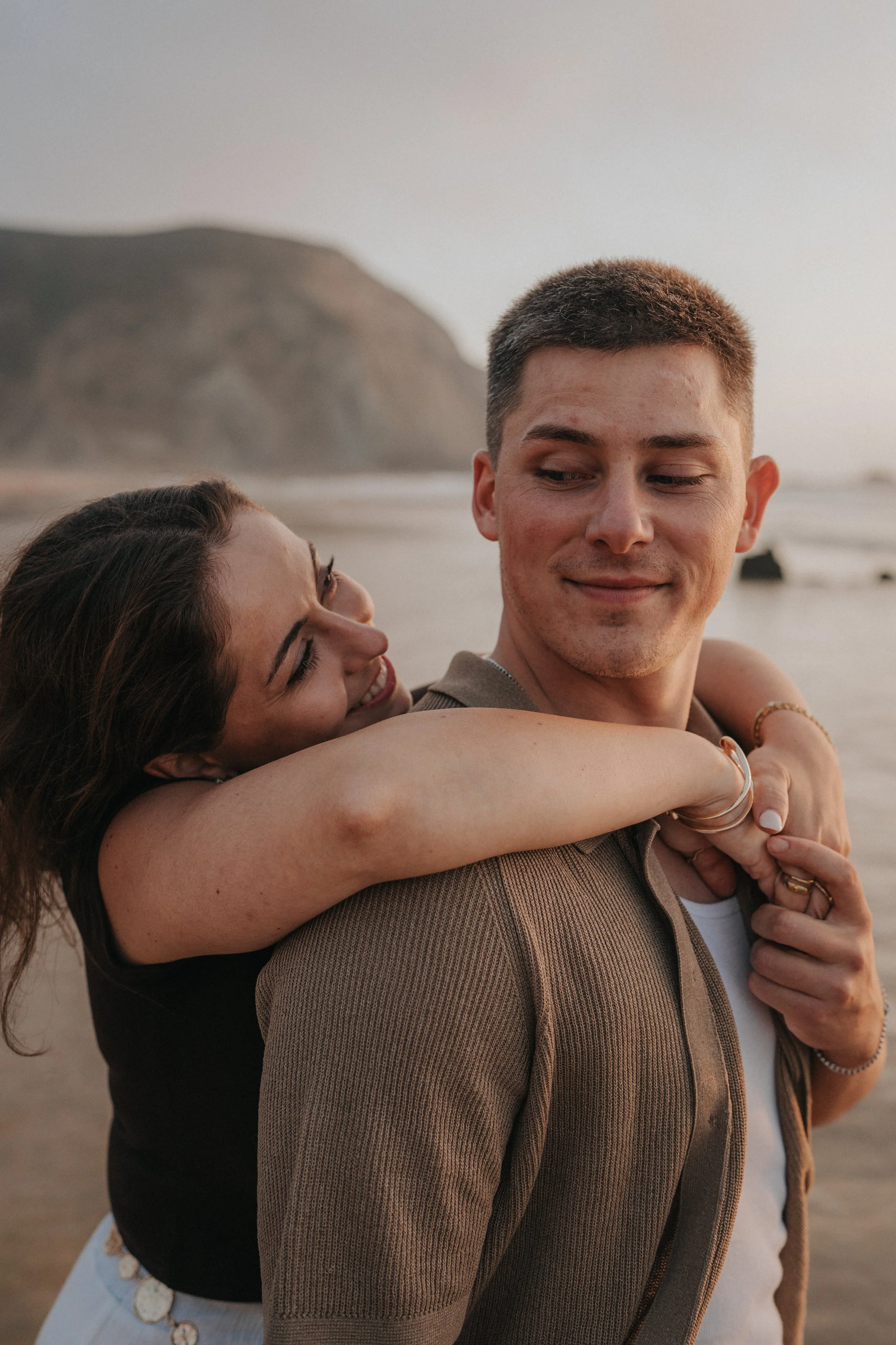 A smiling woman hugging a man at the beach during sunset, with cliffs in the background.