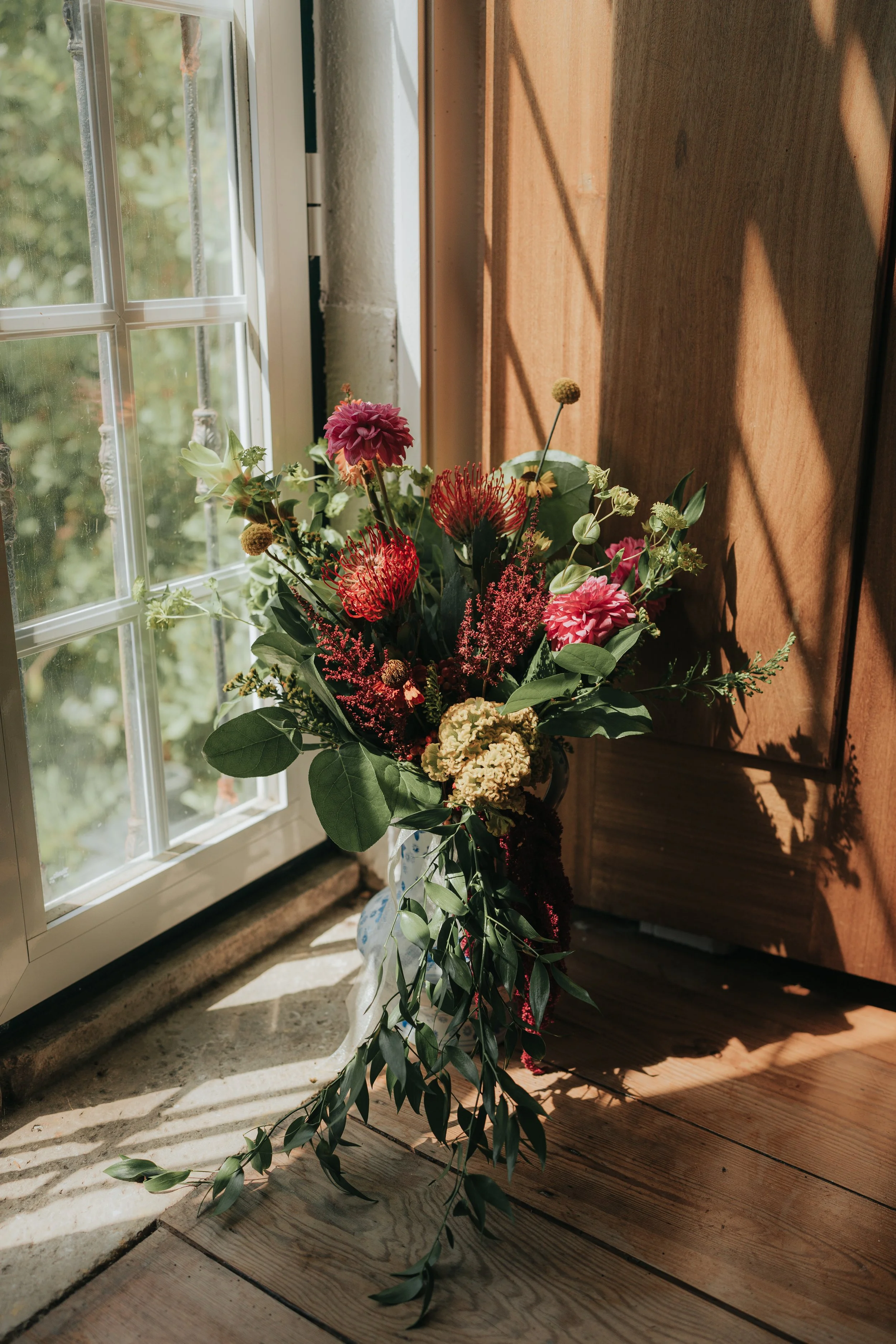 A bouquet of pink, red, and cream flowers with green leaves sitting on a wooden floor next to a window with sunlight casting shadows.