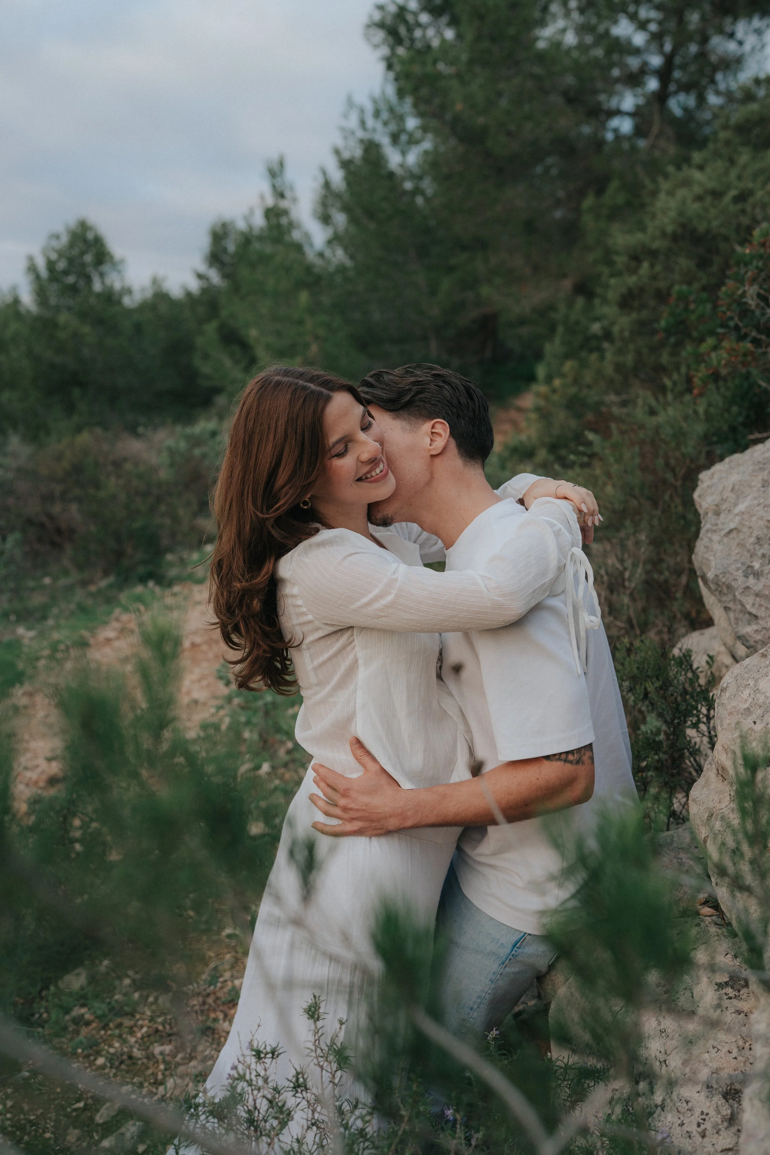 A young woman with long, wavy brown hair and a young man with dark hair and a tattoo on his arm are hugging outdoors in a natural setting with green trees and rocks, smiling and enjoying each other's company.