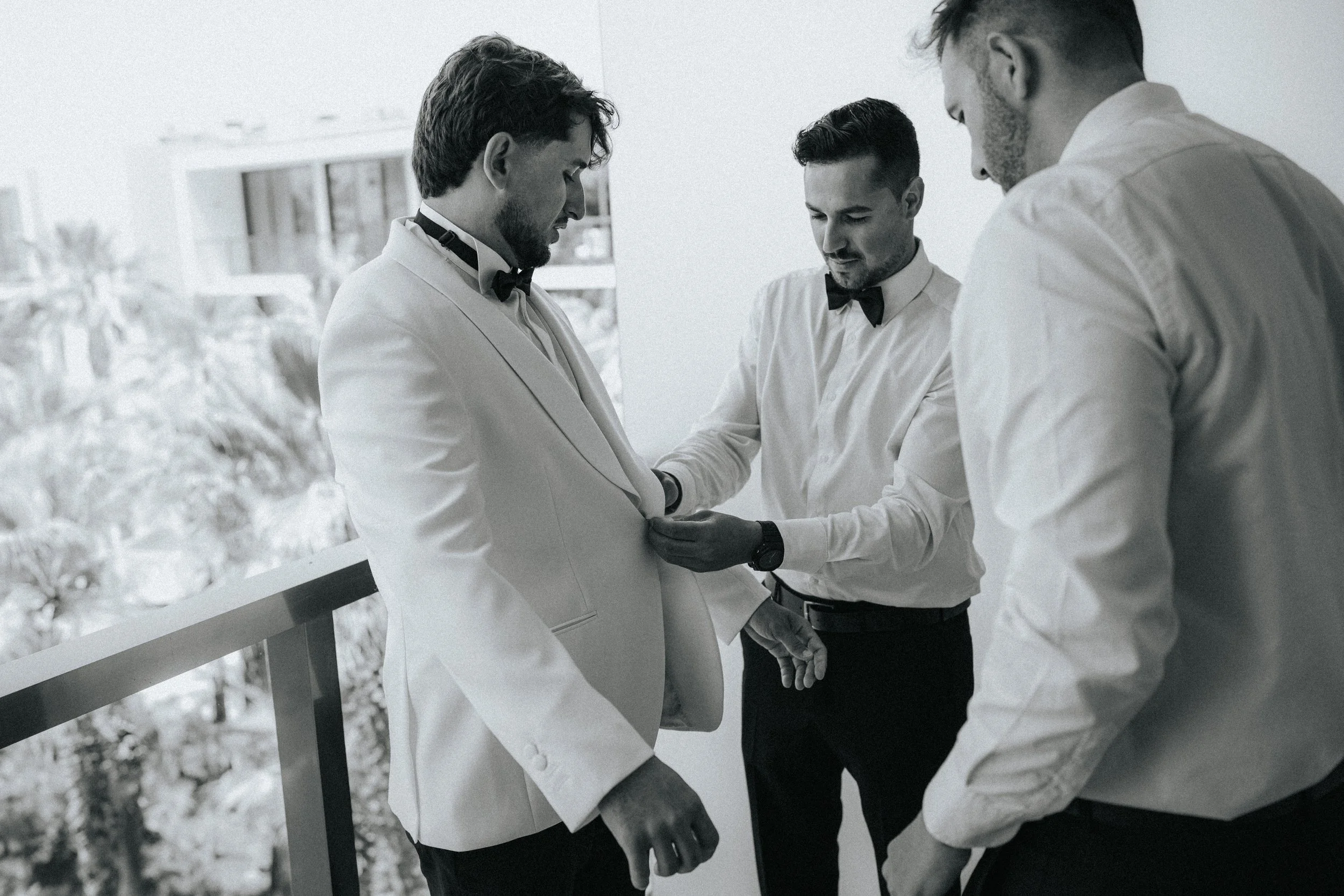 Three men dressed in white tuxedos with black bow ties are standing outdoors on a snowy day, with one man placing a ring on another's finger as part of a wedding ceremony.