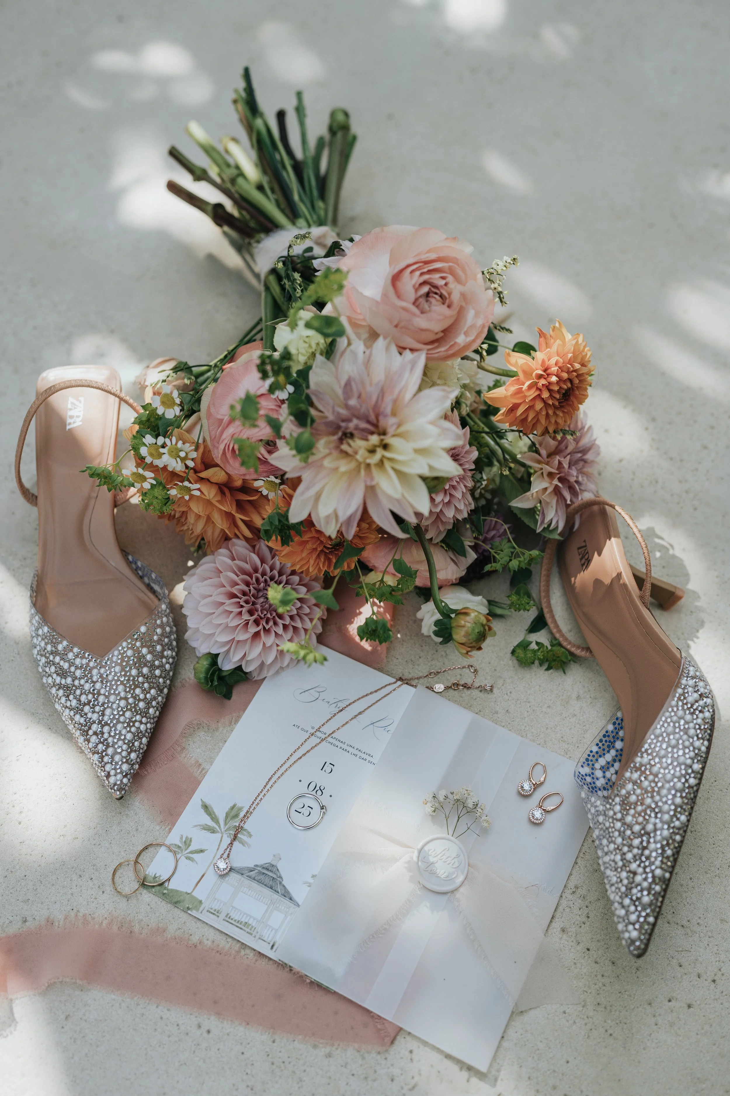 A bridal bouquet of pink and peach flowers, surrounded by pearl-embellished shoes, jewelry, and wedding invitations on a concrete surface with dappled sunlight.