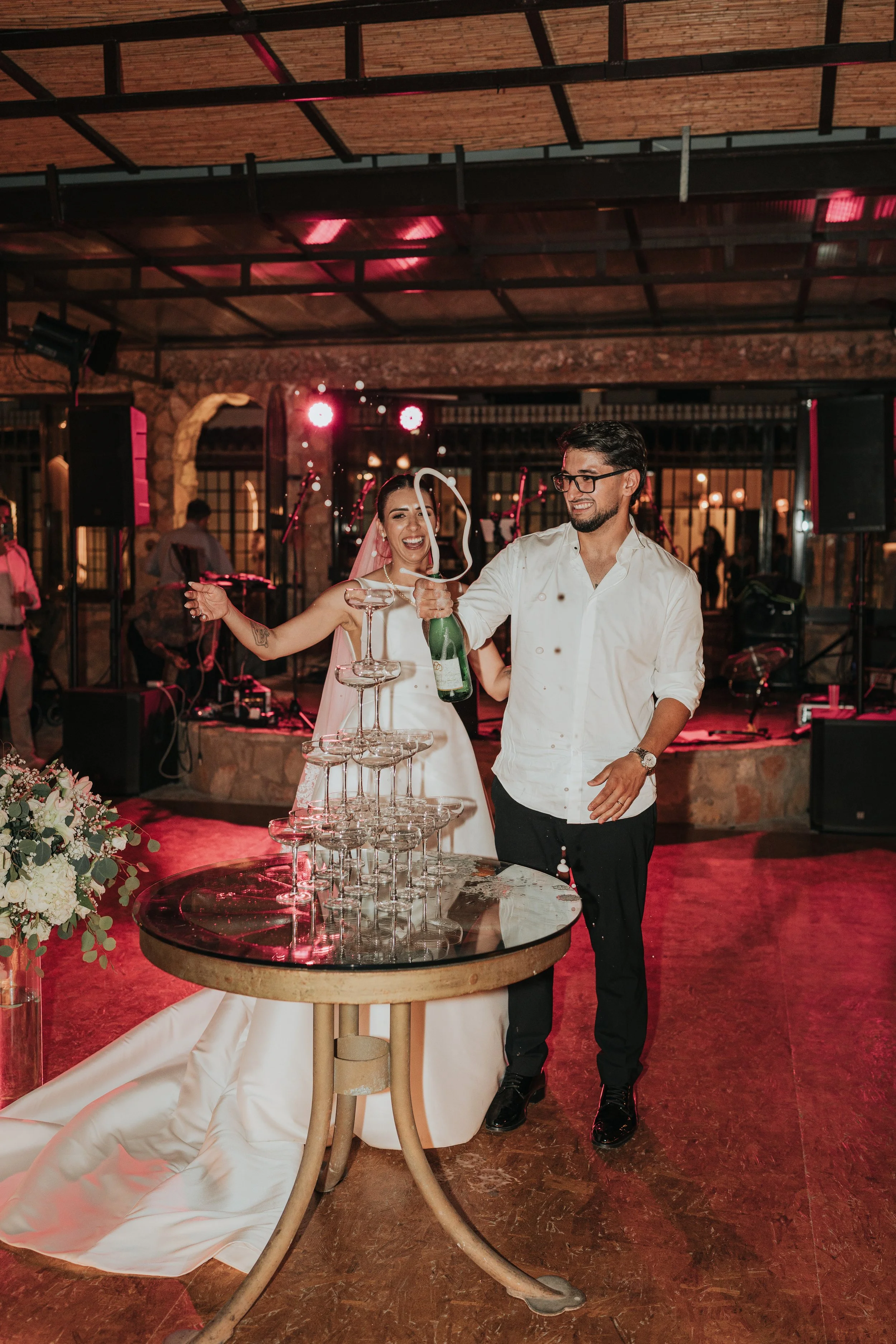 A newlywed couple celebrates at their wedding reception, with the bride in a white wedding dress and the groom in a white shirt and black pants. They are smiling and pouring champagne into a pyramid of glasses on a round table.