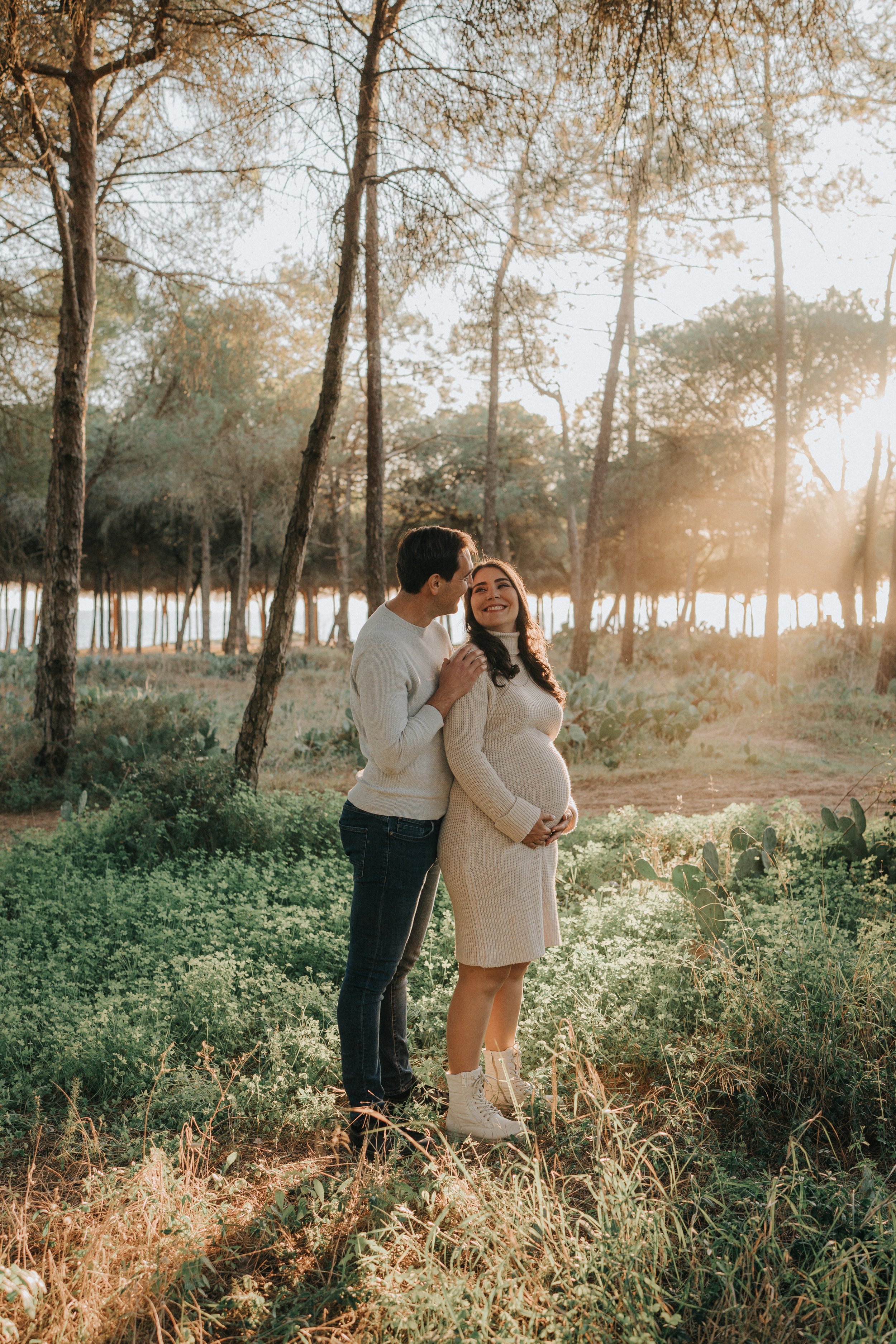 A couple, with the woman visibly pregnant, standing in a forested area during sunset. The man is whispering to the woman, who is smiling. The woman is wearing a beige sweater dress and white boots, while the man is wearing a white sweater and jeans.