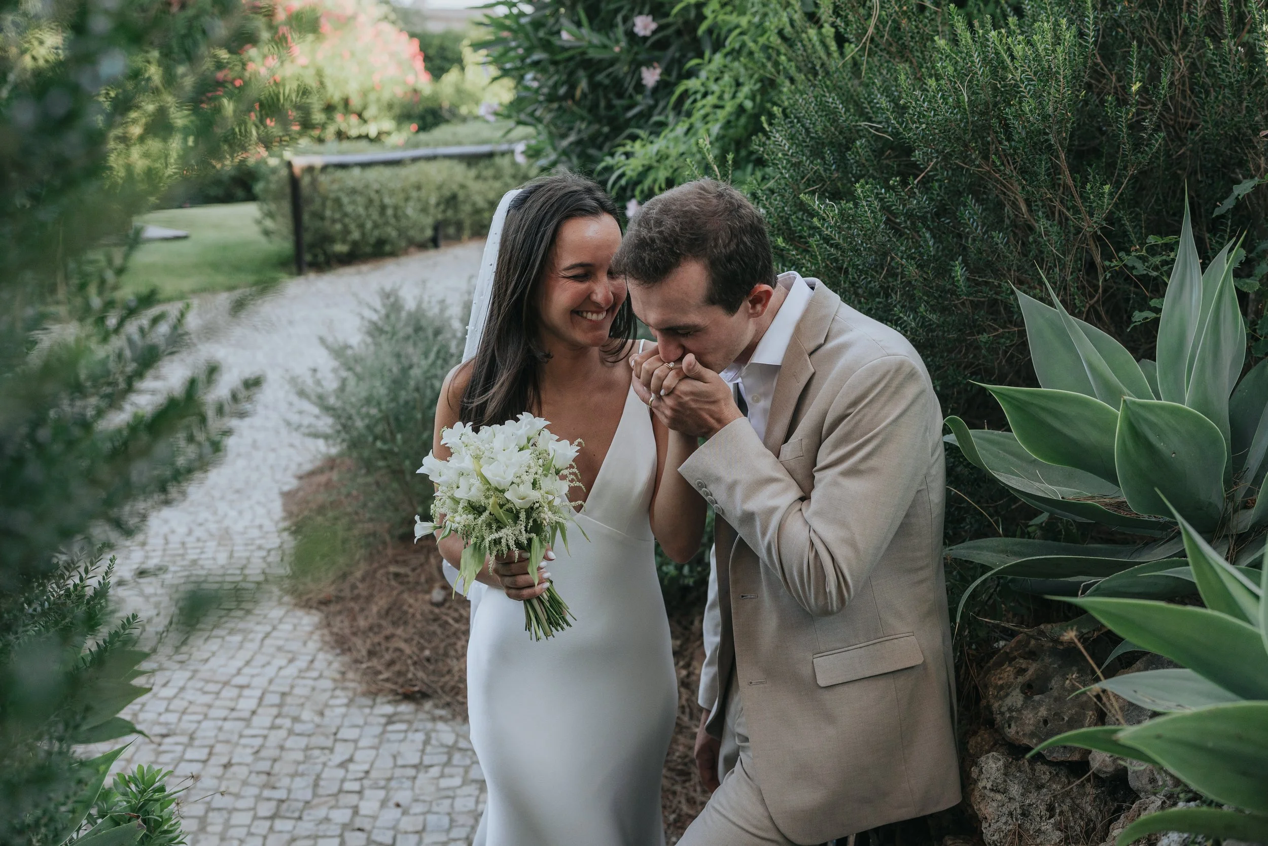 A bride and groom share a romantic moment outdoors, with the groom kissing the bride's hand while she holds a bouquet of white flowers, surrounded by lush greenery.