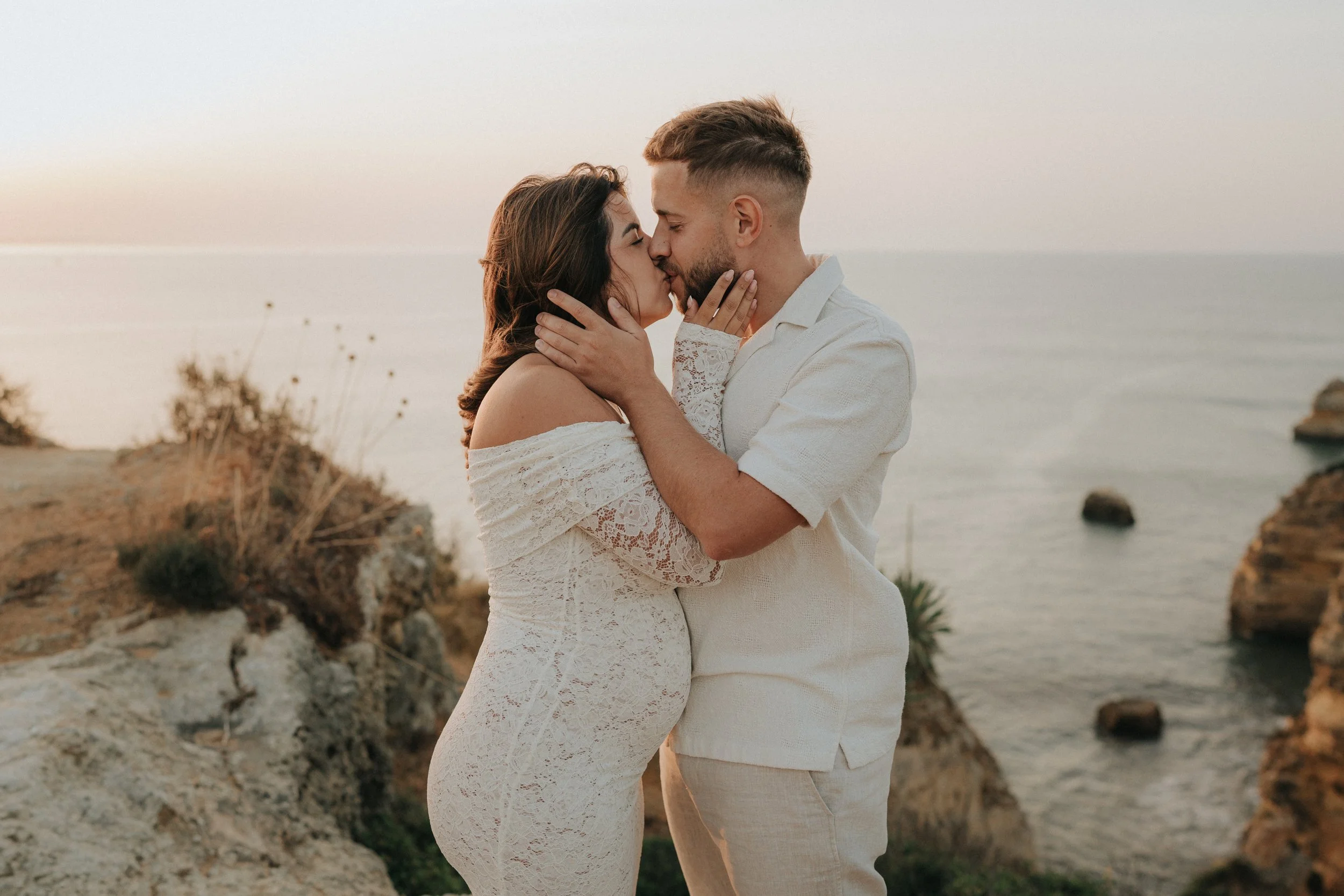 A couple wearing white lace clothing sharing a kiss on a rocky coastline at sunset.