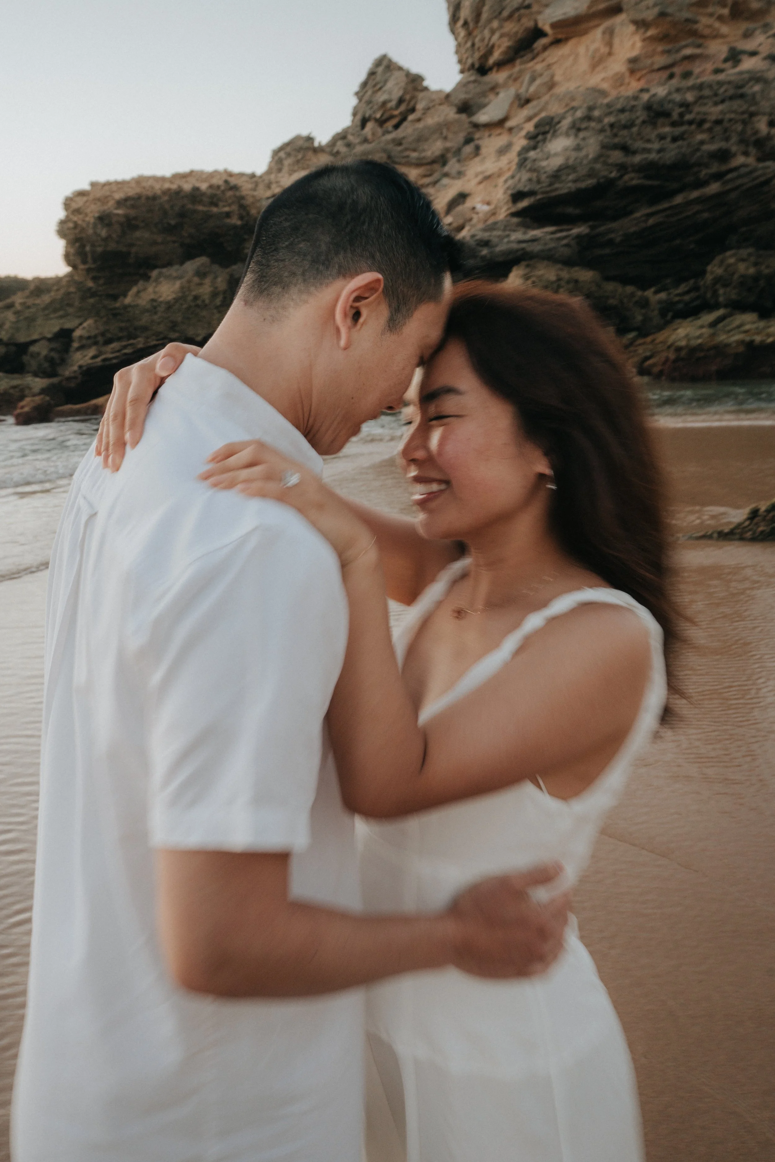 Couple embracing on a beach with large rocky cliffs in the background, smiling and close with foreheads touching.