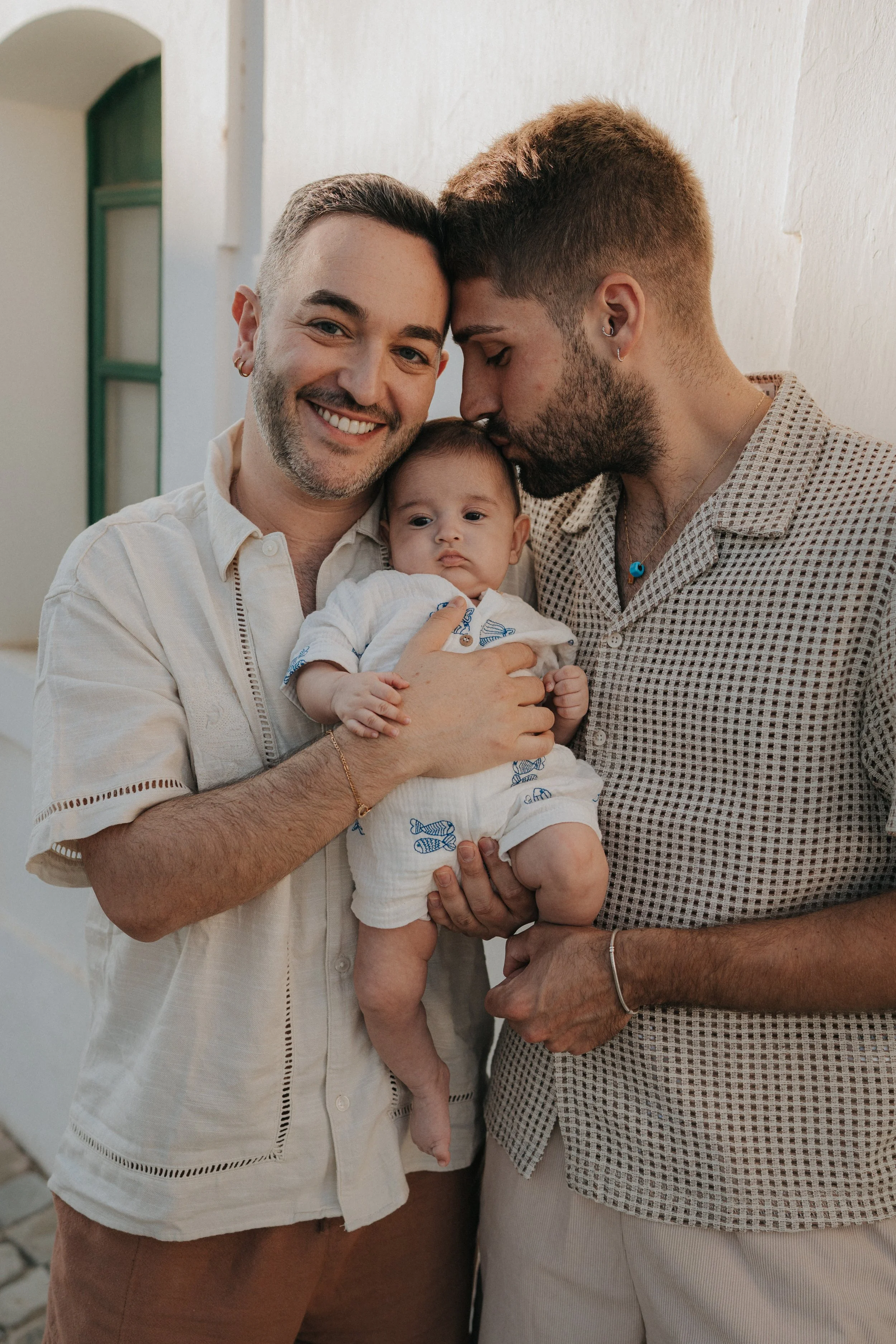 Two men holding a baby, smiling and leaning their foreheads together against a white wall in a well-lit room.