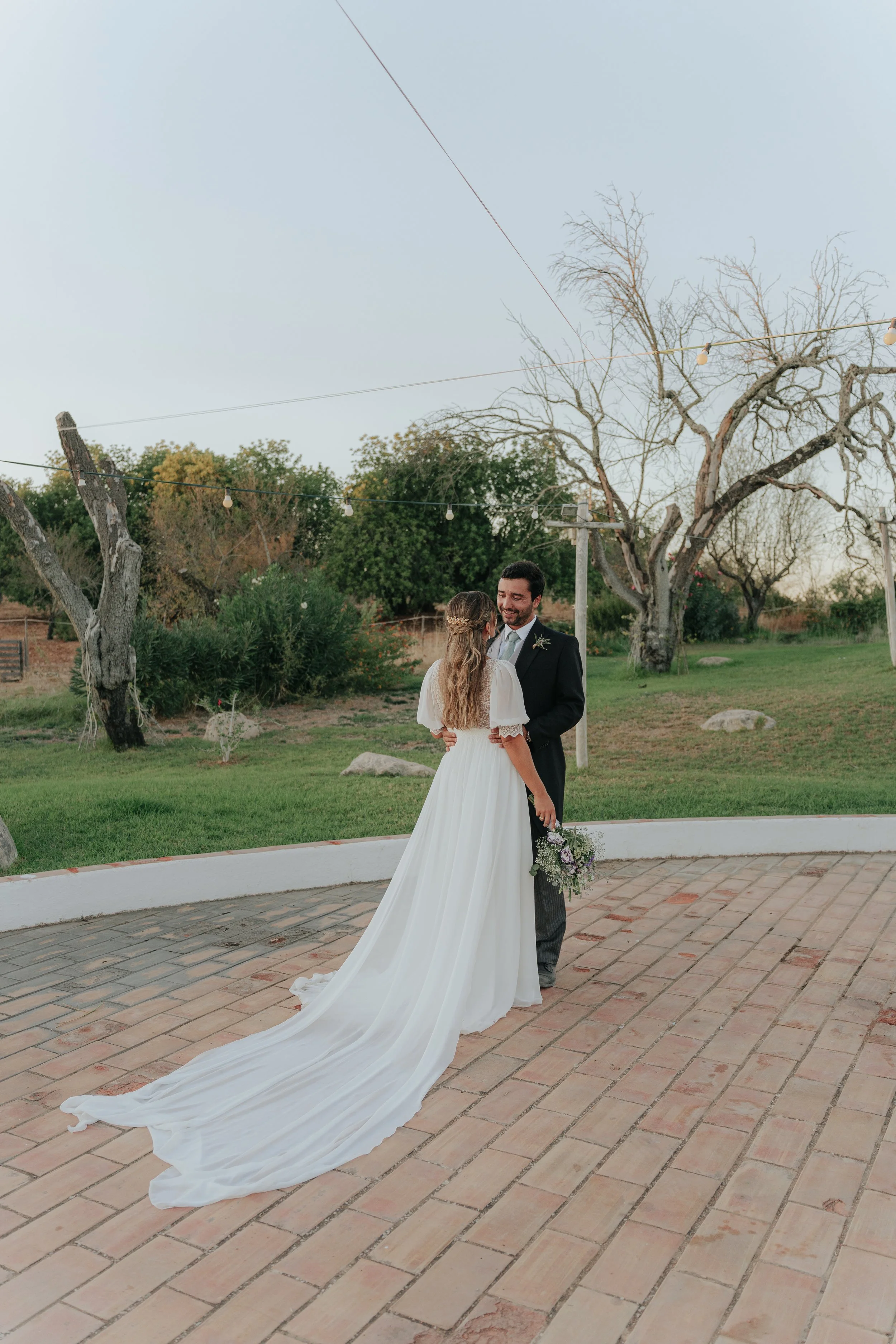 A bride and groom are smiling and embracing during their wedding ceremony outdoors, with trees and string lights in the background.