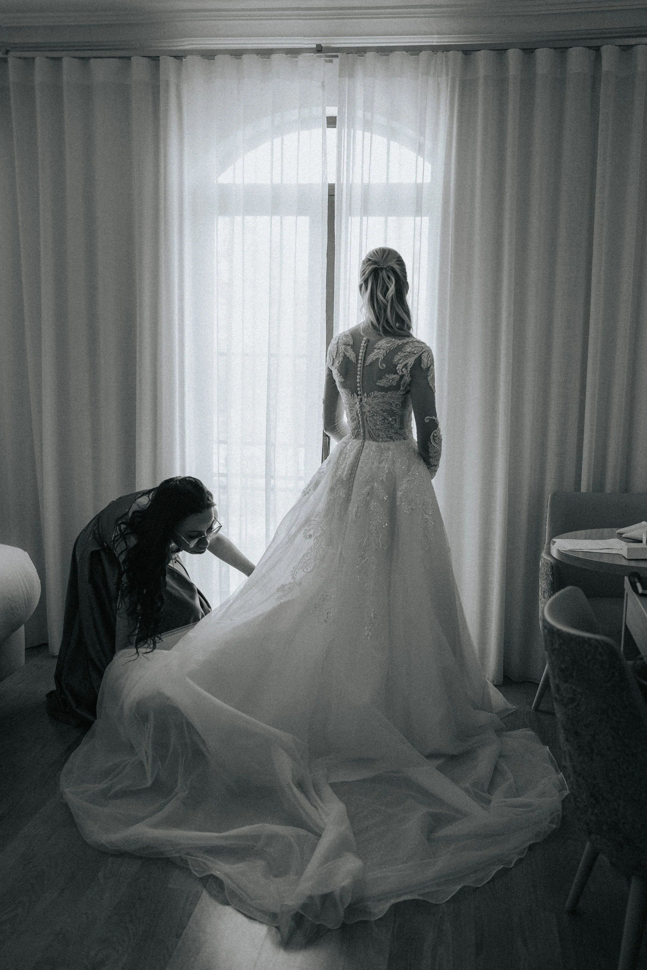 Black-and-white photo of a woman in an ornate wedding dress, standing in front of a window with sheer curtains, while another woman adjusts the gown.