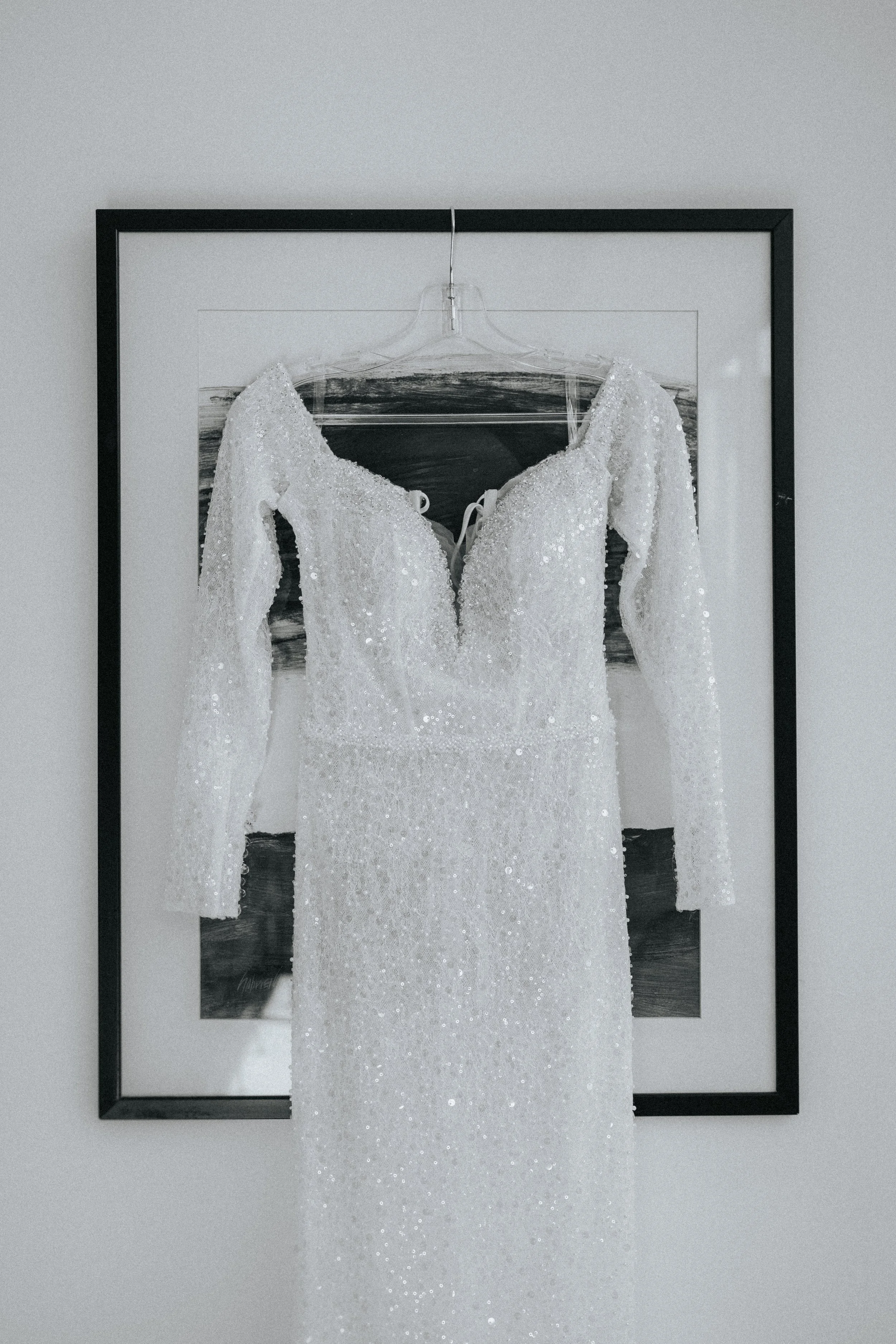 A wedding dress hanging on a clear plastic hanger inside a black picture frame, displayed against a white wall with a small picture of a beach scene in the background.