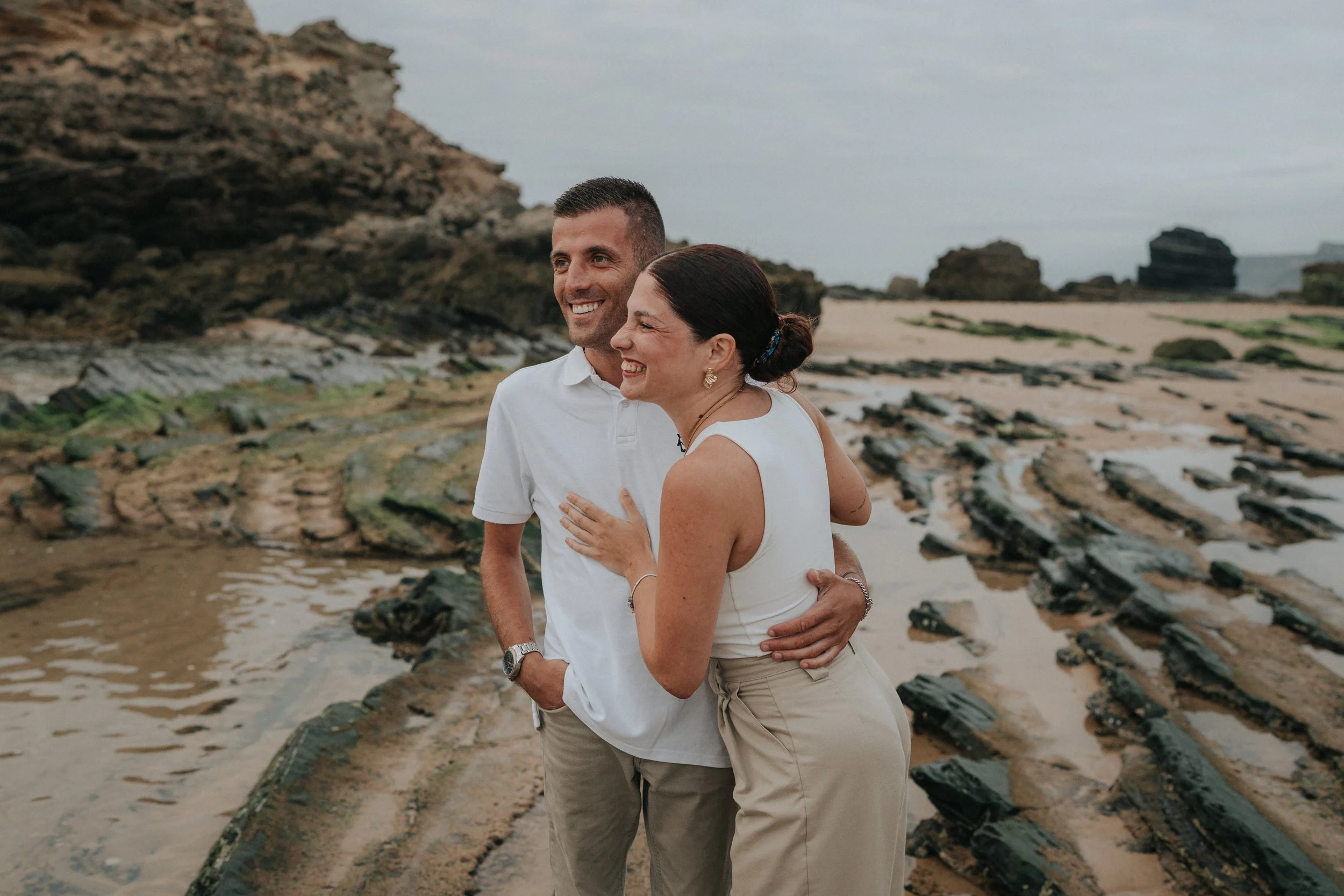 A smiling couple standing on the beach with rocky formations in the background, embracing each other.