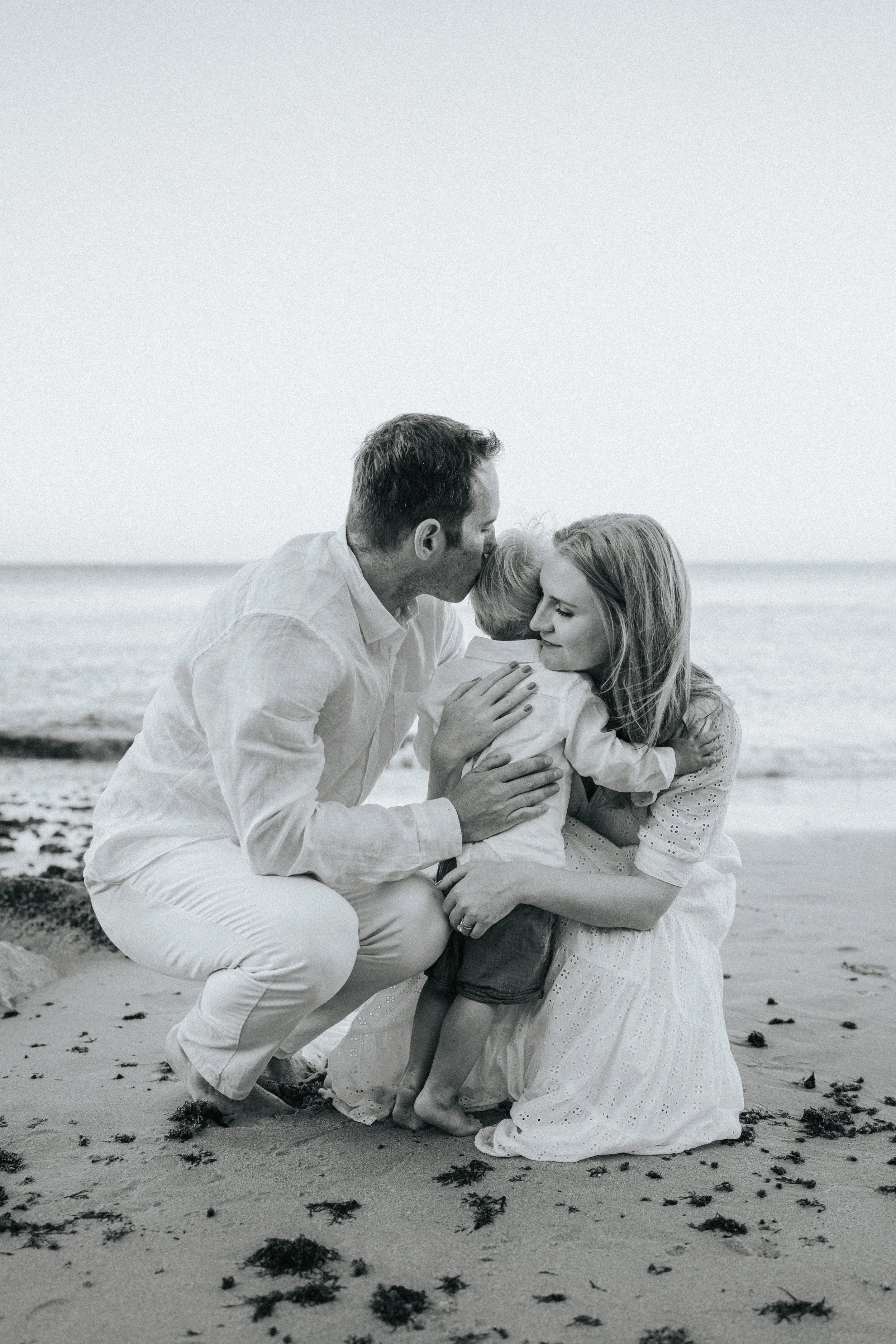 A black and white photo of a family at the beach. The man is giving a kiss on the head to a woman who is hugging a young girl. The family is sitting on the sand with seaweed around them, and the ocean is in the background.
