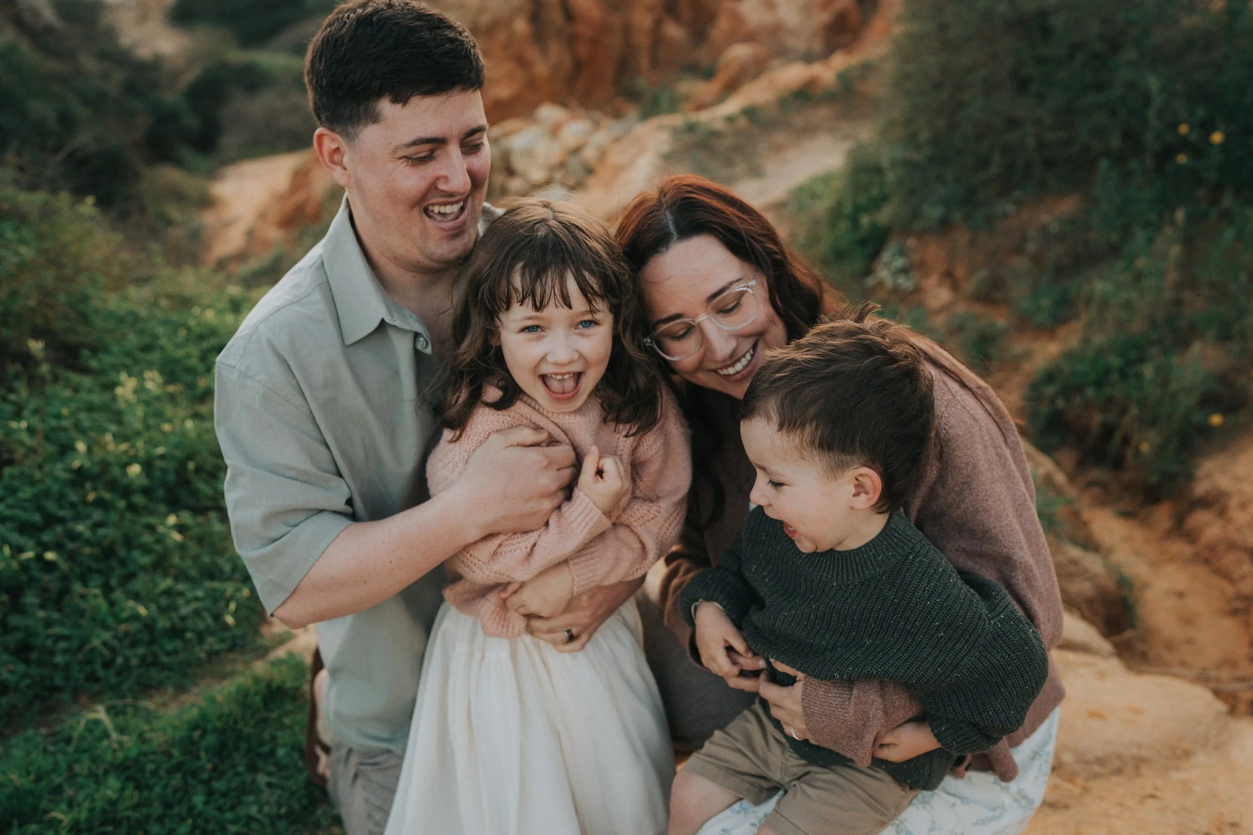 A joyful family group hugging outdoors in a natural setting with greenery and rocky terrain in the background, smiling and laughing together.