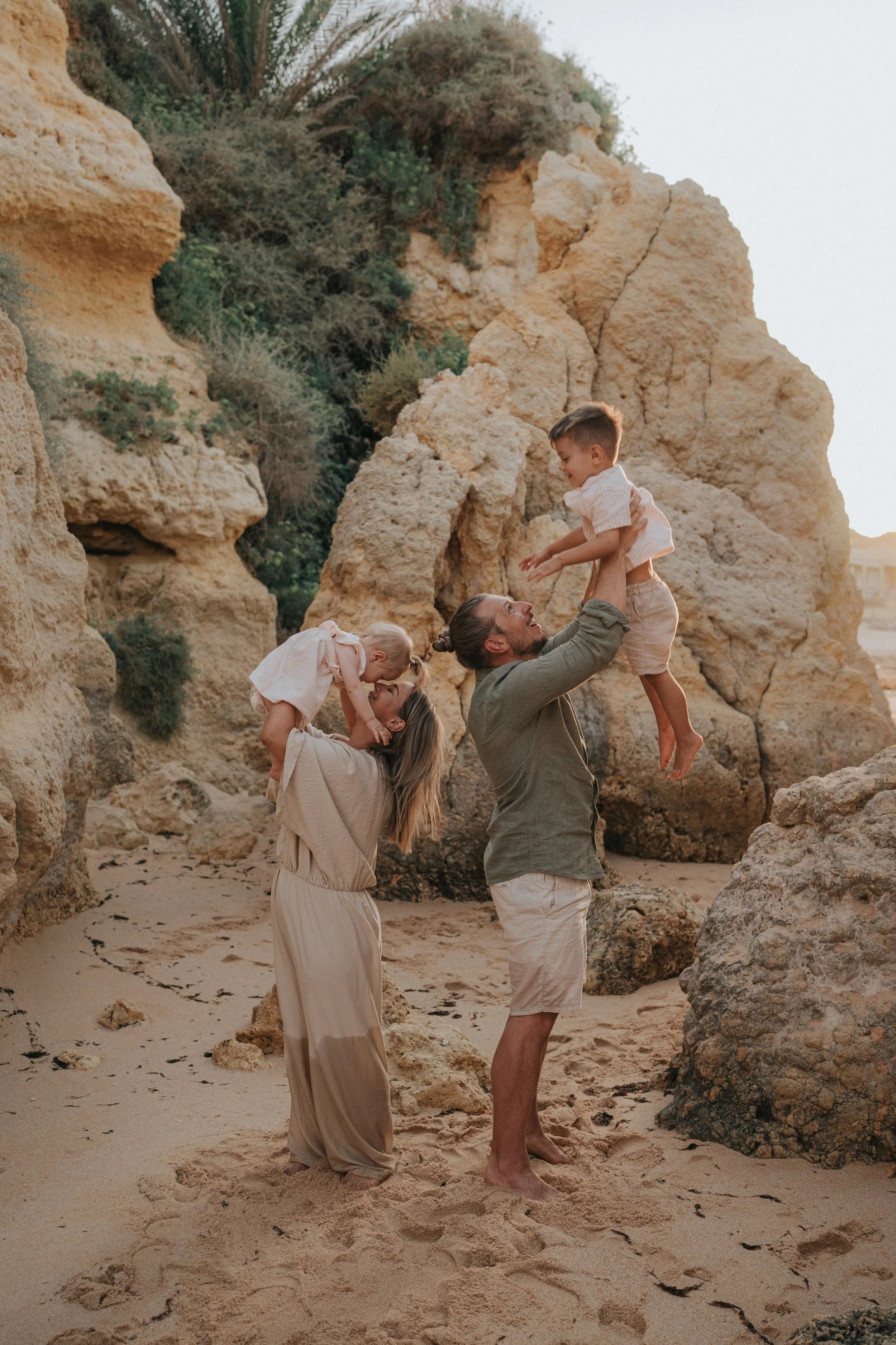 A family of four enjoying time together on a rocky beach with beige rocks and sandy ground. The father lifts a young boy in the air while the mother holds a small girl, all smiling and engaging playfully.