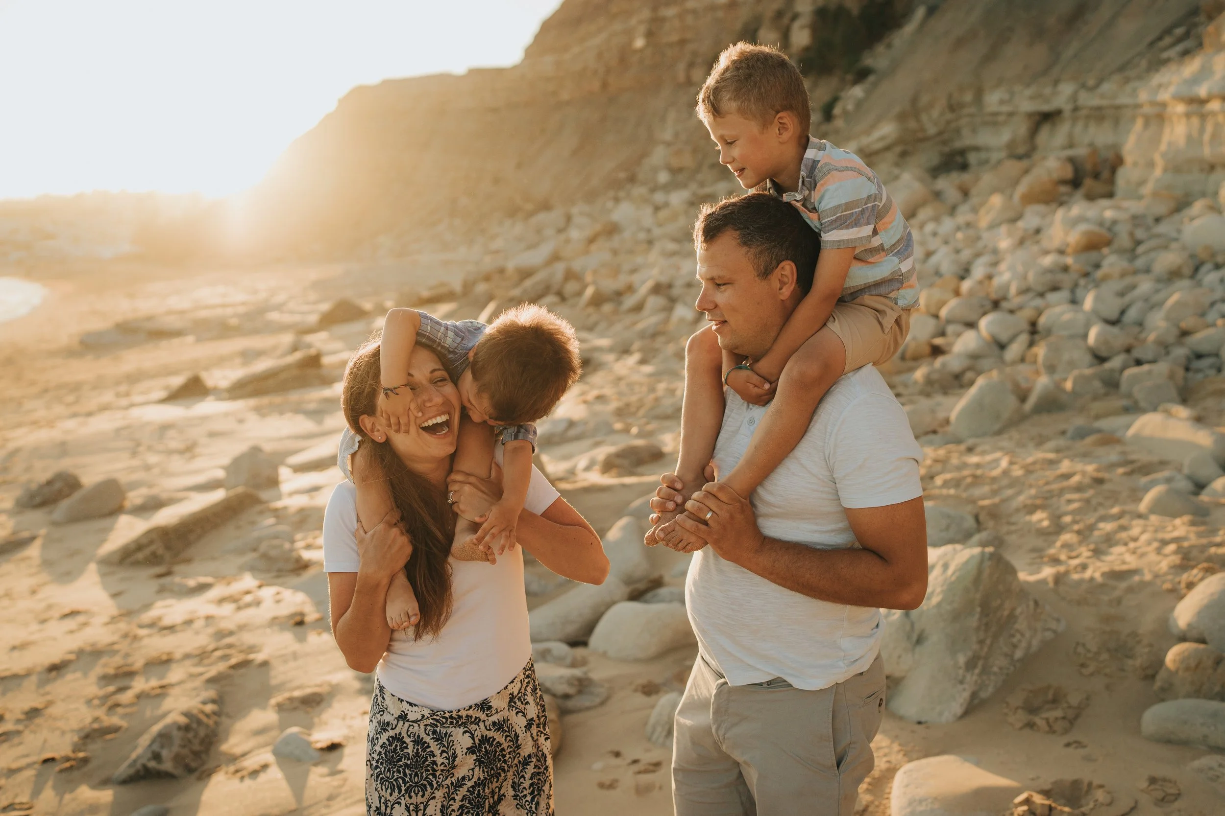 A family of four enjoying the beach. The mother, wearing a white top and patterned skirt, is holding her young son on her shoulder. The father, in a white shirt and khaki pants, carries another son on his shoulders. The children are smiling and the f