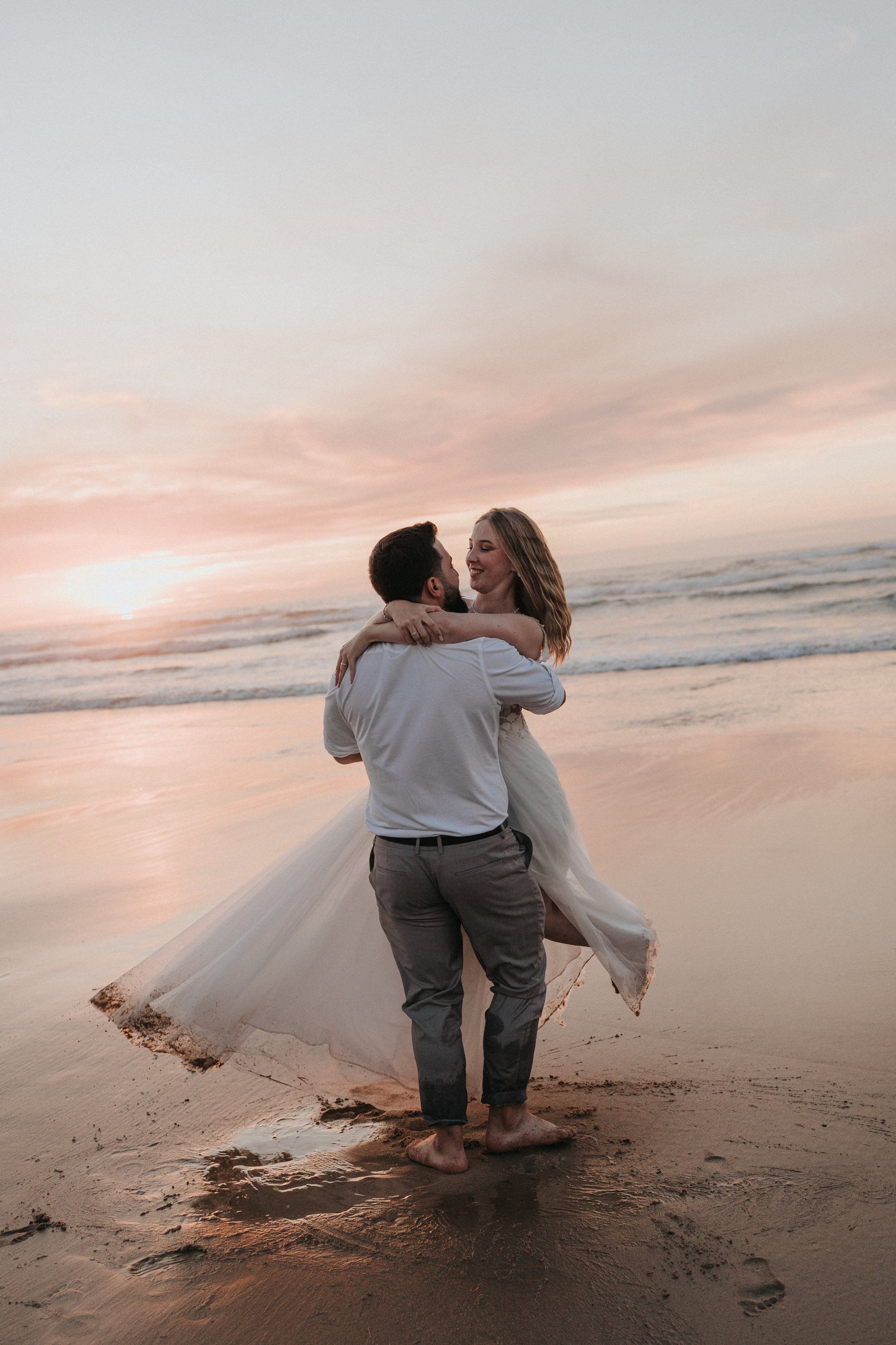 A couple is seen embracing on a beach during sunset, with the man lifting the woman, who is wearing a wedding dress, in his arms. They are barefoot on the sand, and the sky has soft pink and orange hues.