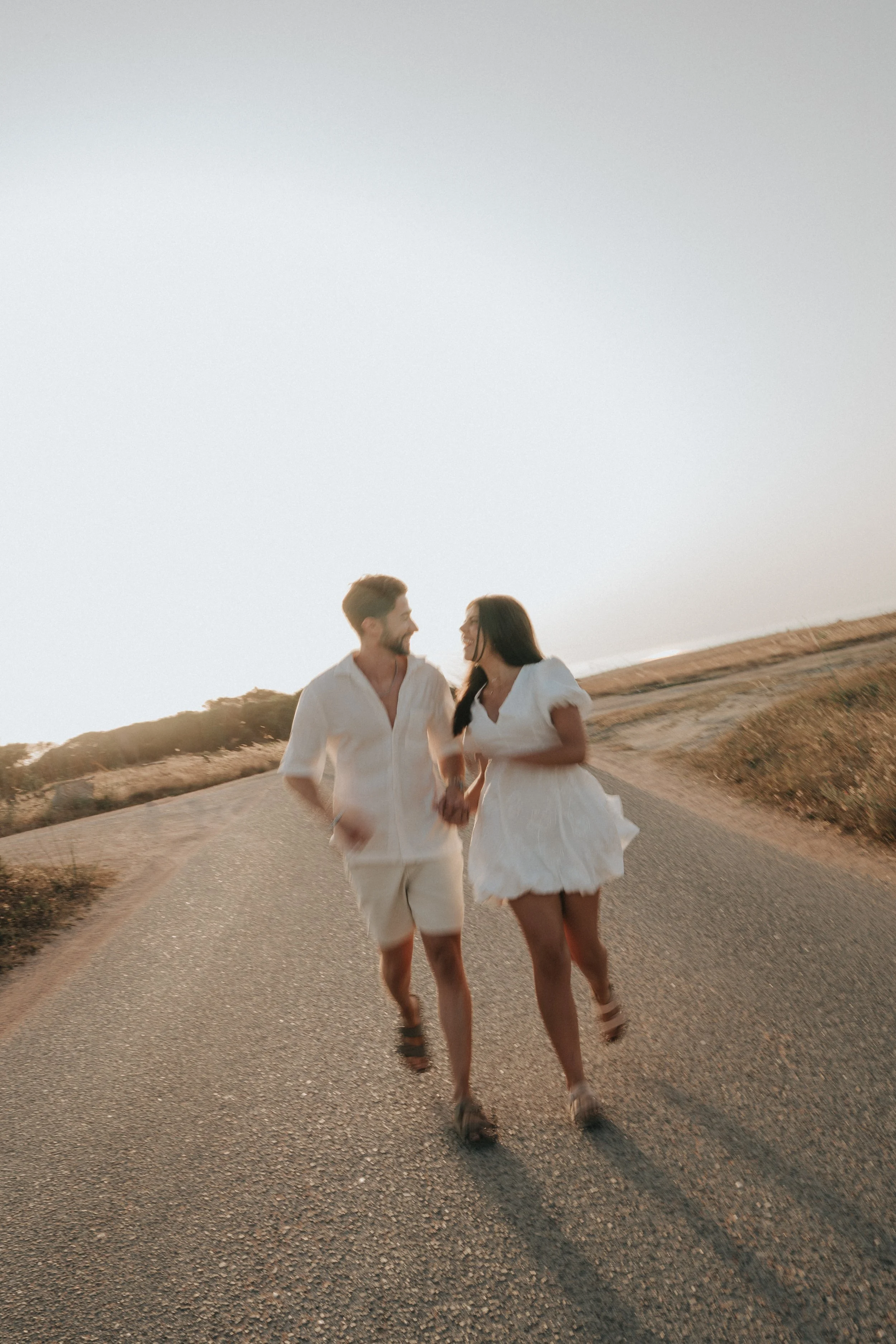 A couple holding hands and walking along a paved road in a desert landscape during sunset, both smiling and looking at each other, dressed in light-colored summer clothing.