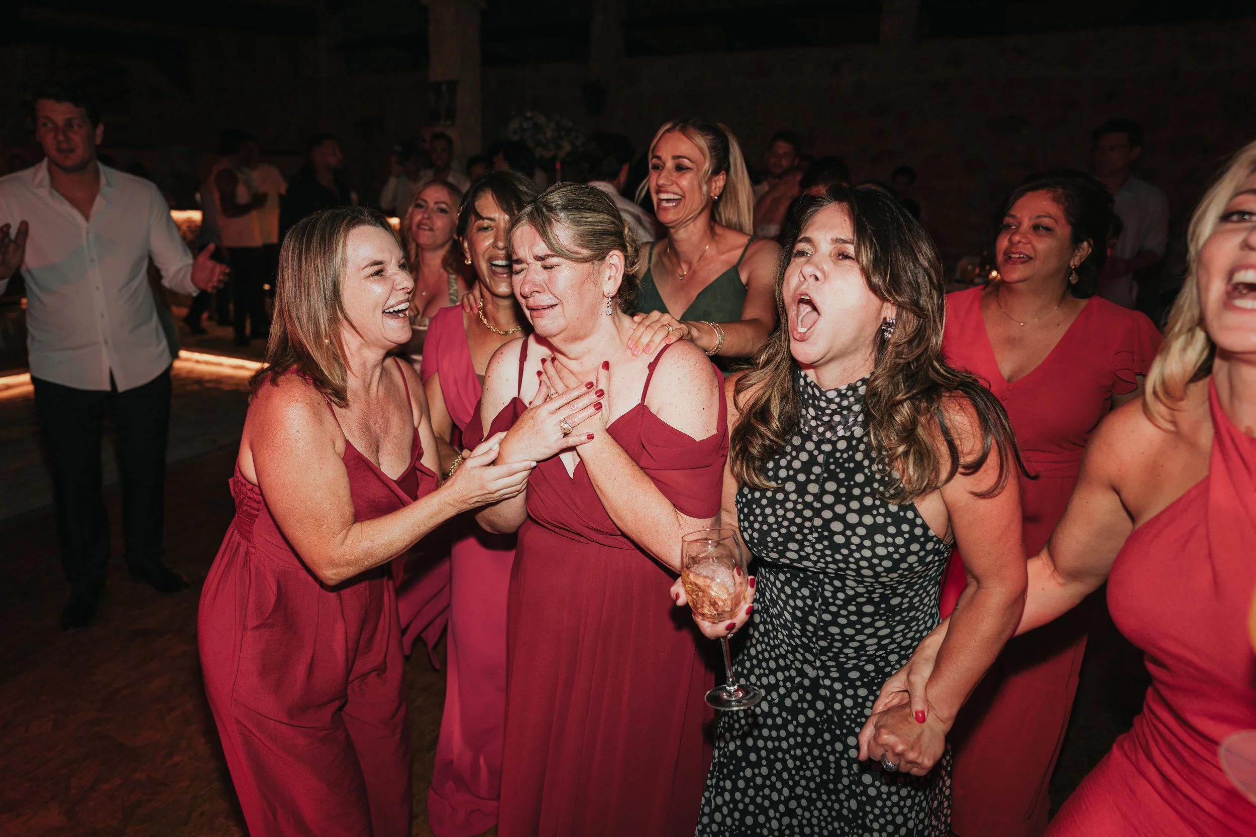 Group of women smiling, laughing, and embracing at a party, some holding drinks, in a dimly lit indoor venue.