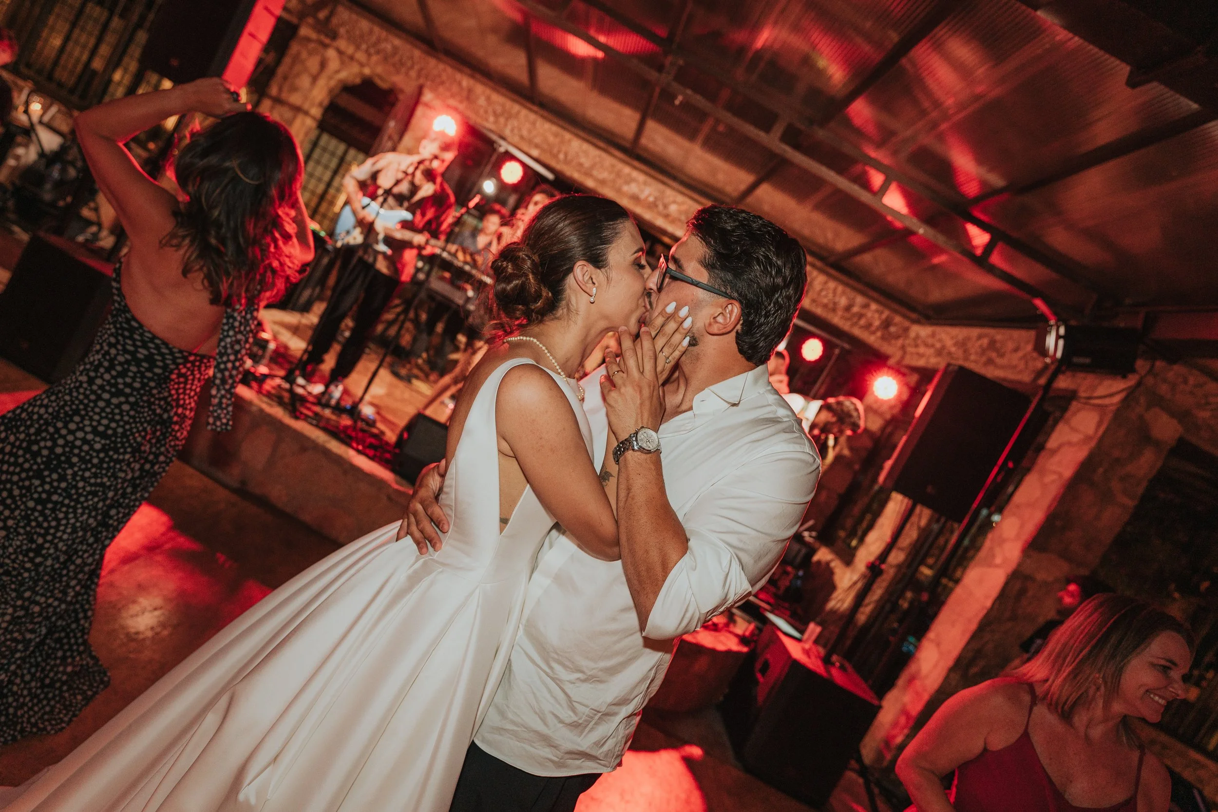 A couple kissing each other on the dance floor at a lively event, with women dancing and a live band playing in the background.