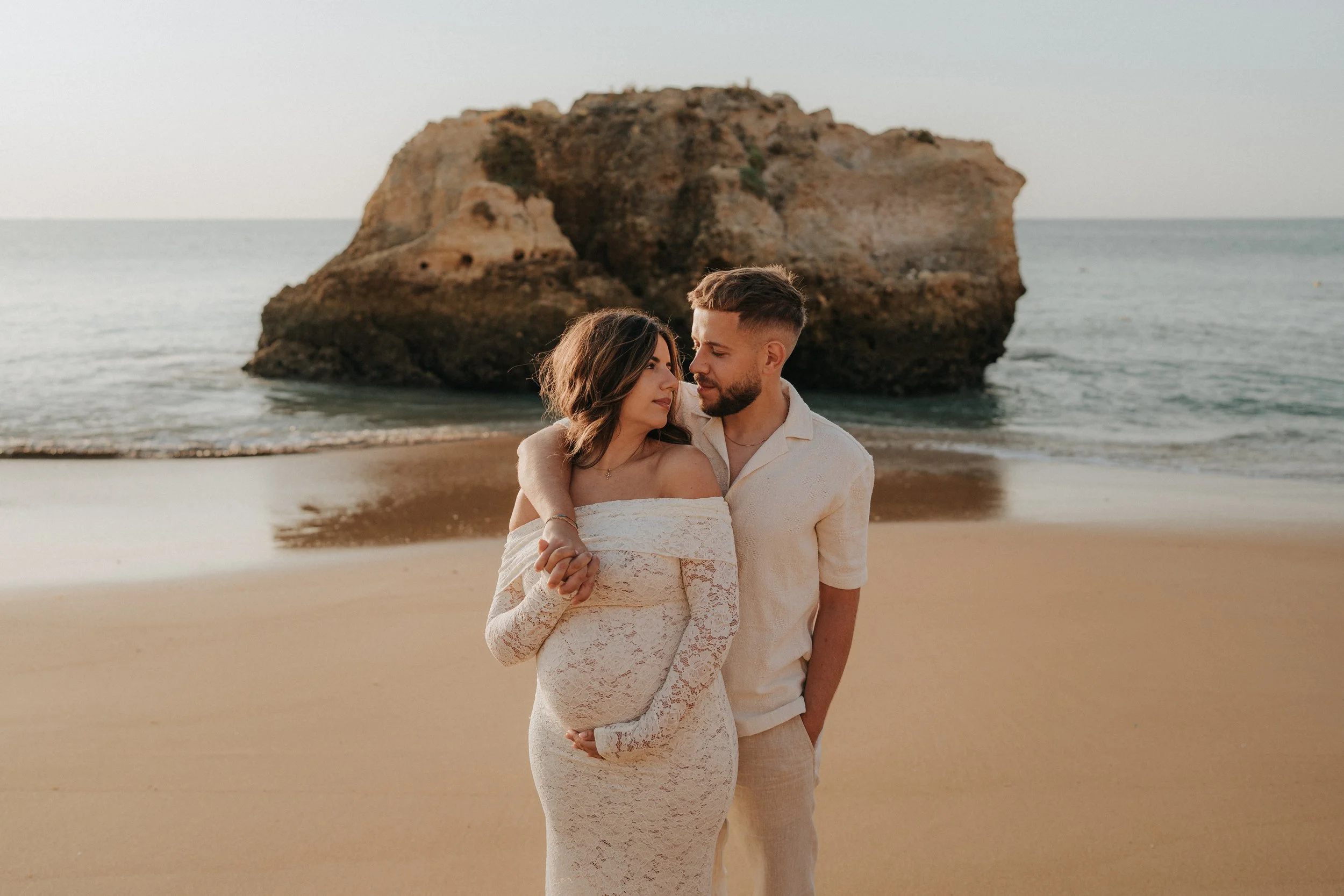 A couple stands on a beach near a large rock formation, embracing each other and looking into each other's eyes during sunset.