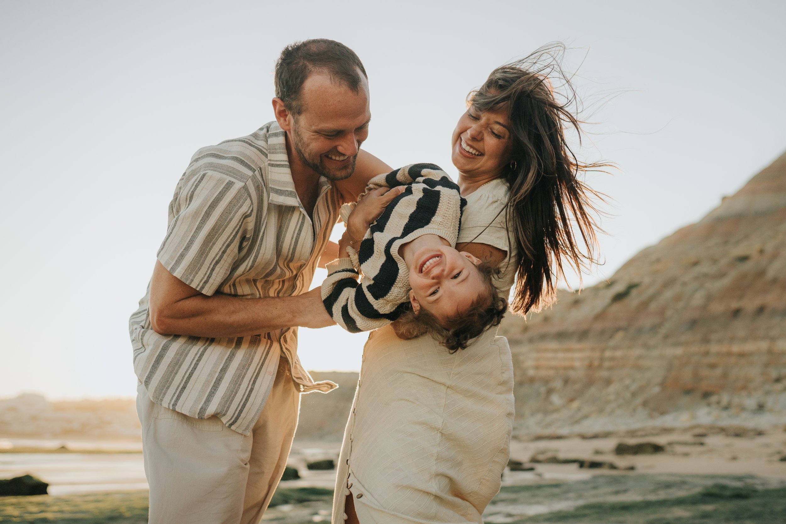 A happy family of three, a man, a woman, and a young boy, playing and having fun together outdoors in a natural setting during the daytime. They are smiling and the boy is being held upside down by the man, with the woman supporting him. The backgrou