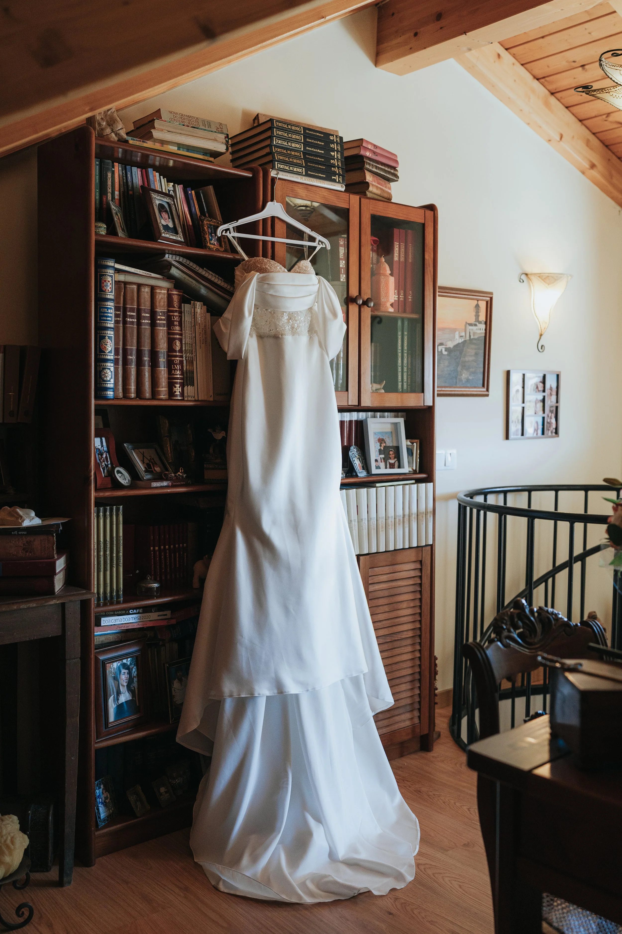 A white wedding dress hanging on a white hanger from a wooden bookshelf in a cozy living room, with framed pictures and books on the shelves.