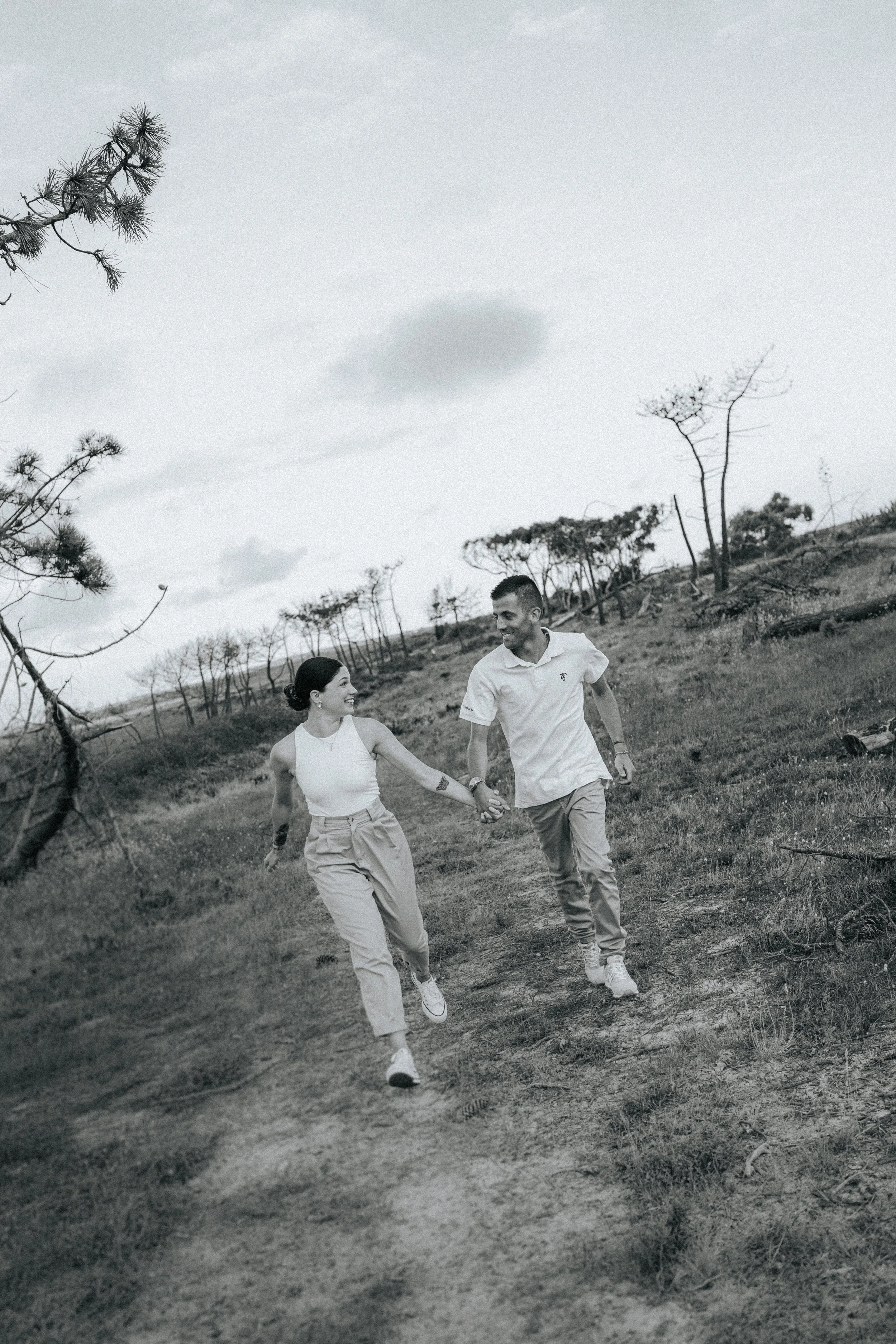 A black and white photo of a man and woman holding hands and walking outdoors on a dirt path with sparse trees around and a cloudy sky.