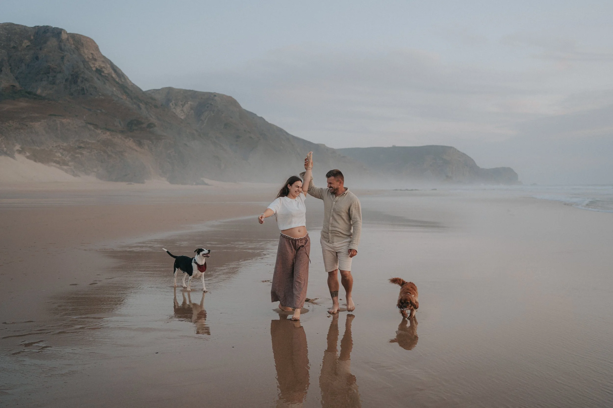 A couple walking on the beach with their two dogs, one black and white and the other brown, near the water's edge with cliffs in the background and the sky partly cloudy.