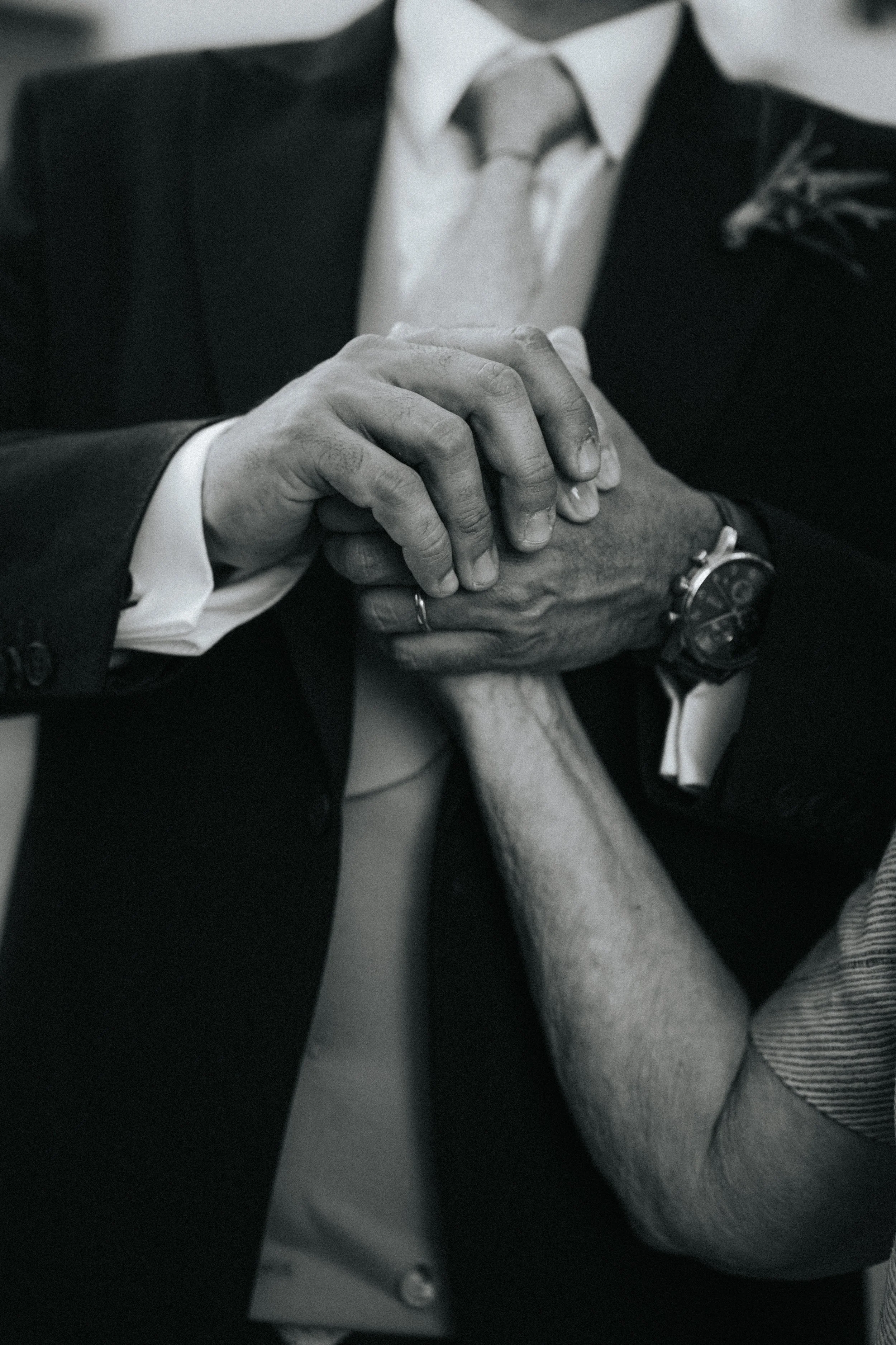 Black and white photo of a person wearing a business suit, holding a smaller person’s hand, who is wearing a watch and a ring.