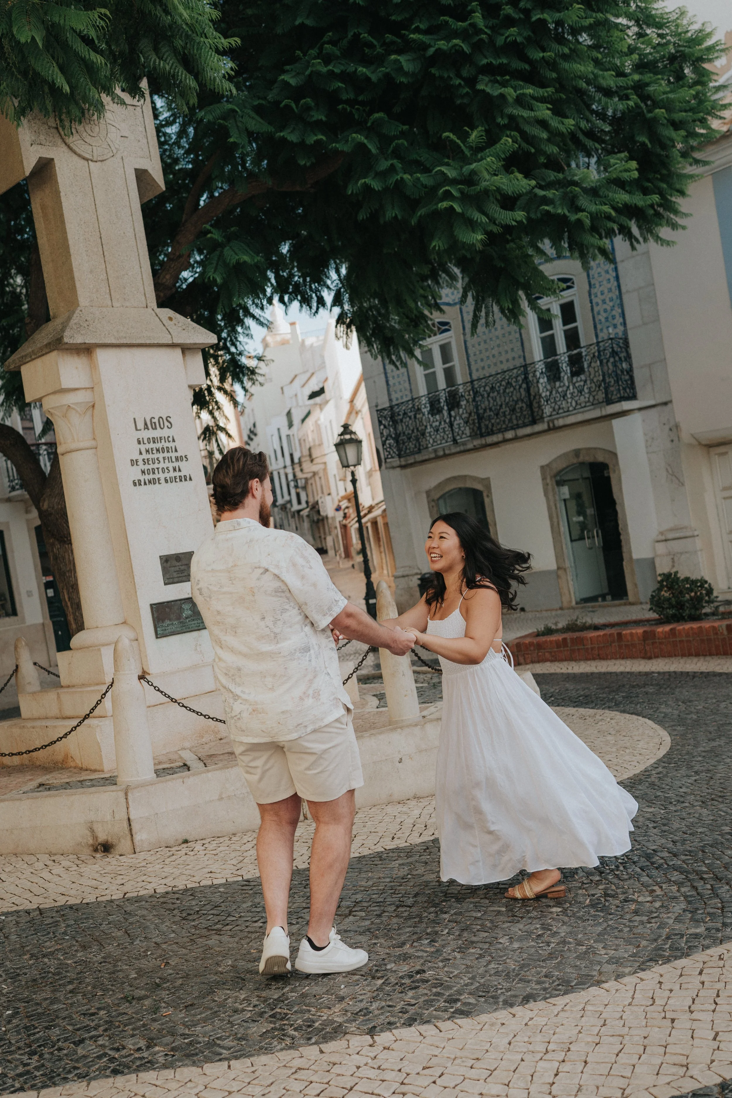 A man and a woman are dancing together outdoors on a cobblestone street, with historic buildings and a large green tree in the background. The man wears a light short-sleeved shirt and shorts, and the woman is in a white dress. They are smiling at ea