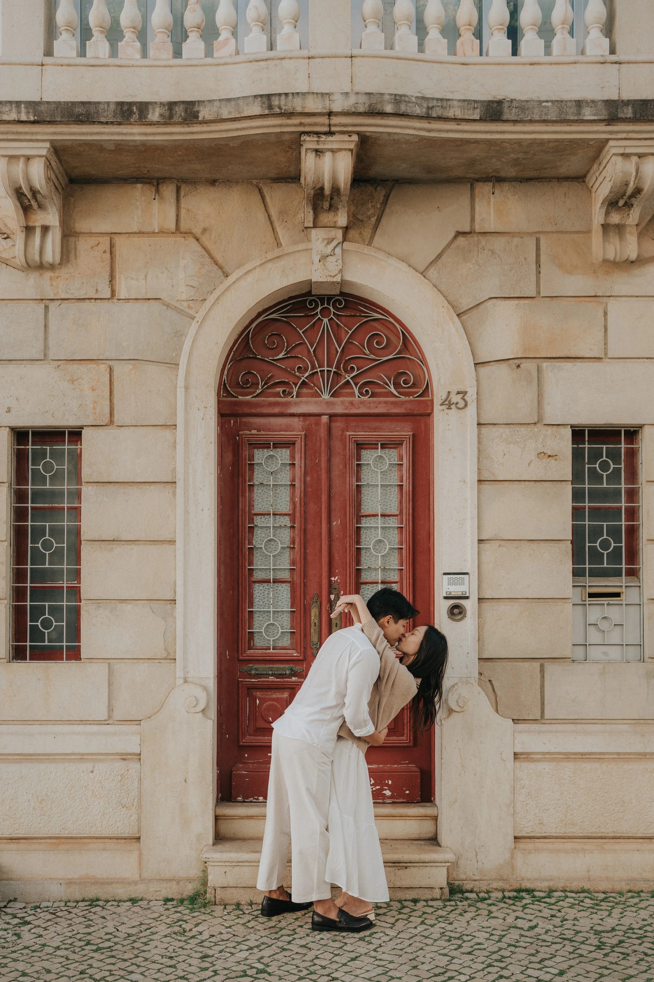 Two people are kissing in front of a vintage-style building with a red door and stone facade. One person is wearing a white outfit, and the other is wearing a beige coat. They are standing on a cobblestone street.