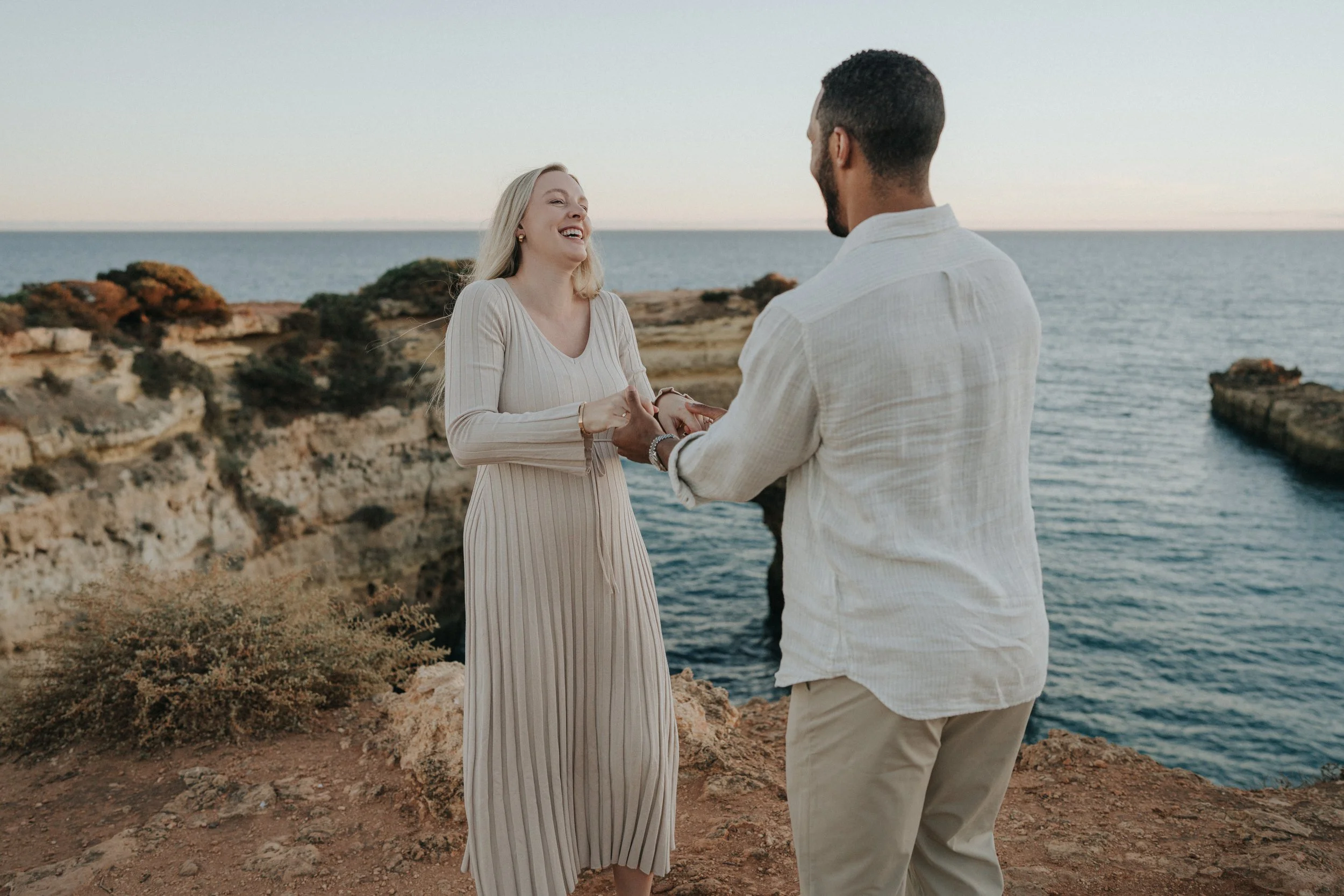 A couple stands on rocky cliffs by the ocean, holding hands and smiling at each other during sunset.