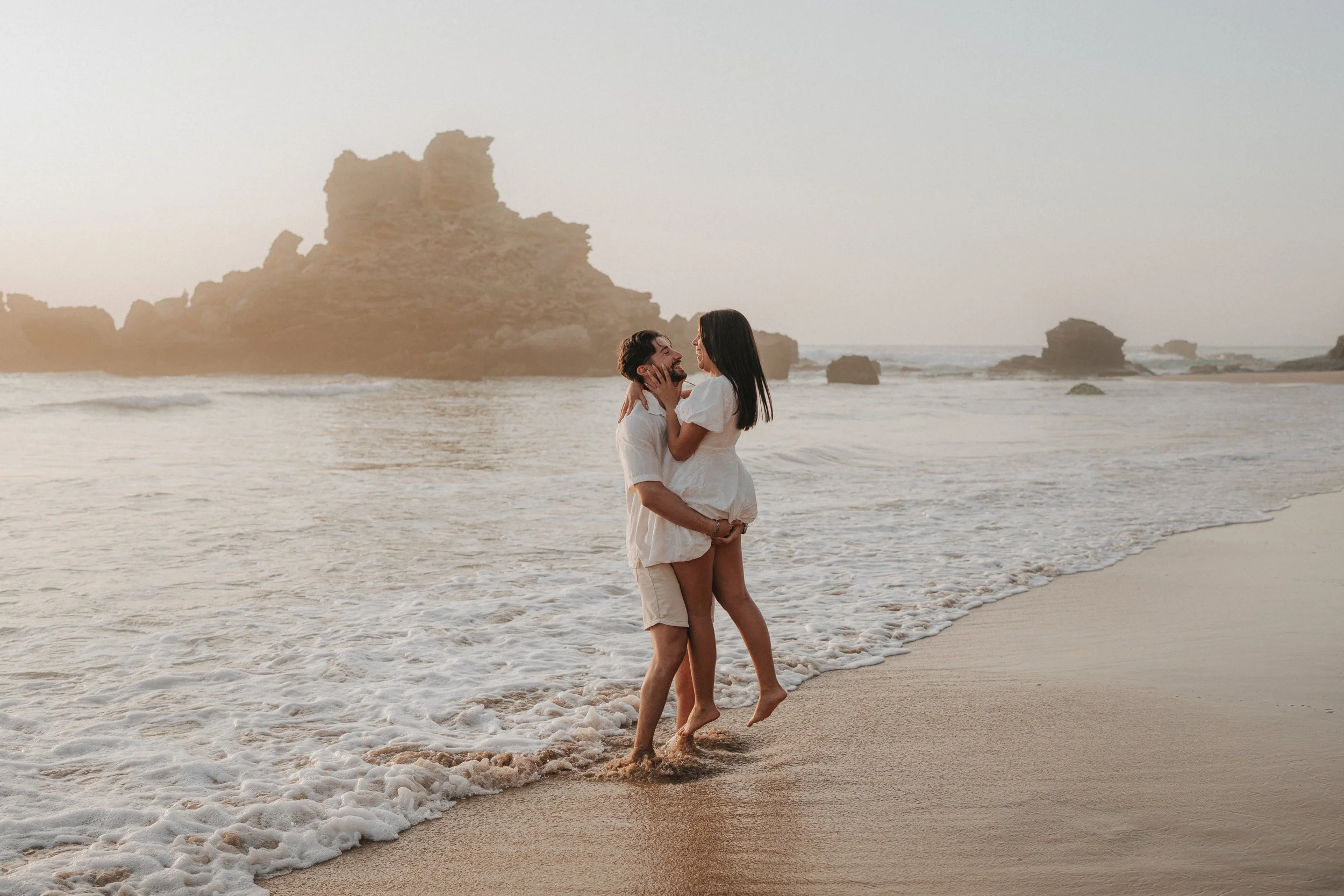 A couple standing in the shallow surf at the beach, smiling and embracing each other with rocky formations in the background during sunset.